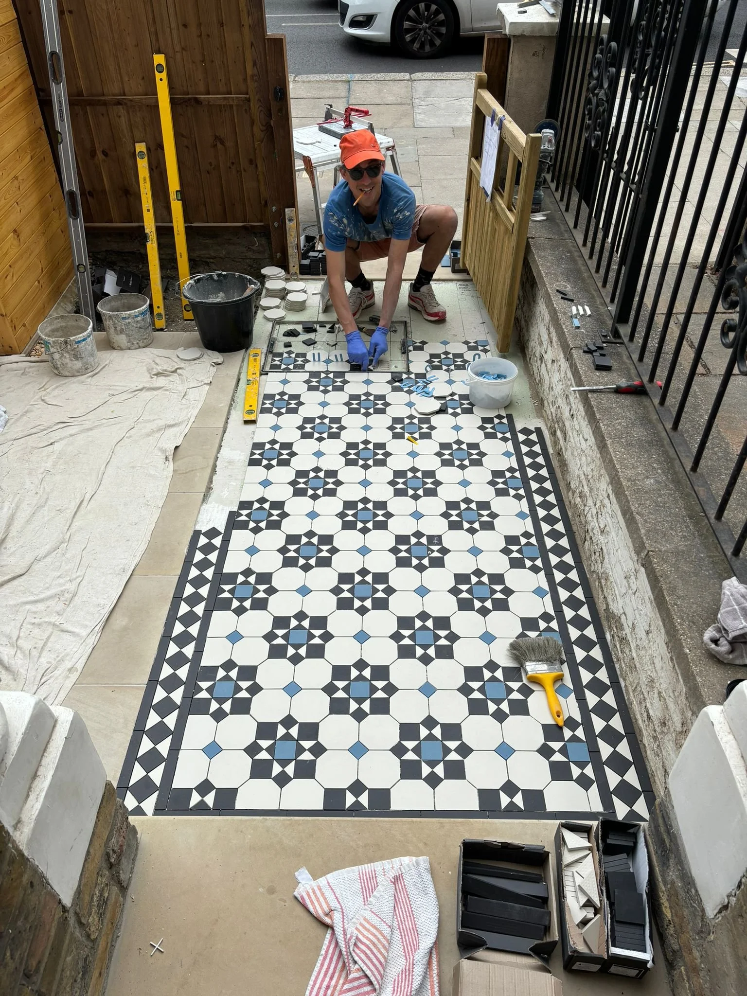 A person installing decorative black and white tiles on a porch floor, surrounded by tools, supplies, and buckets.