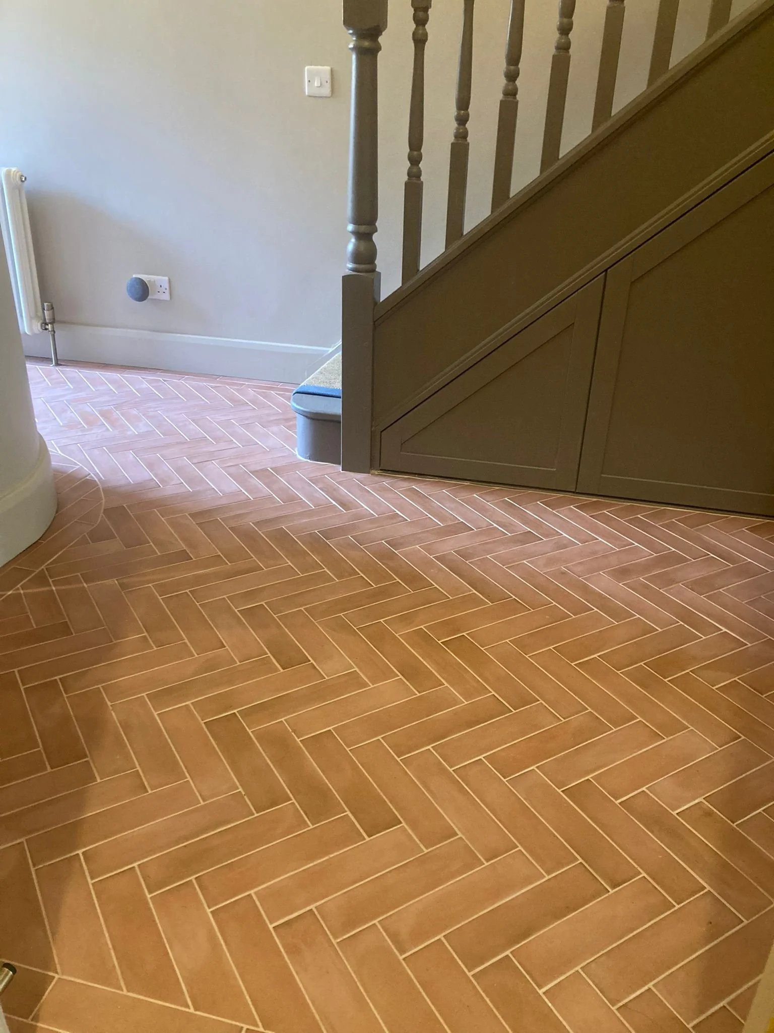 Interior view of a staircase with wooden banister and spindles, a radiator, a wall socket, a small carpeted area at the bottom of the stairs, and a tiled floor in a herringbone pattern.