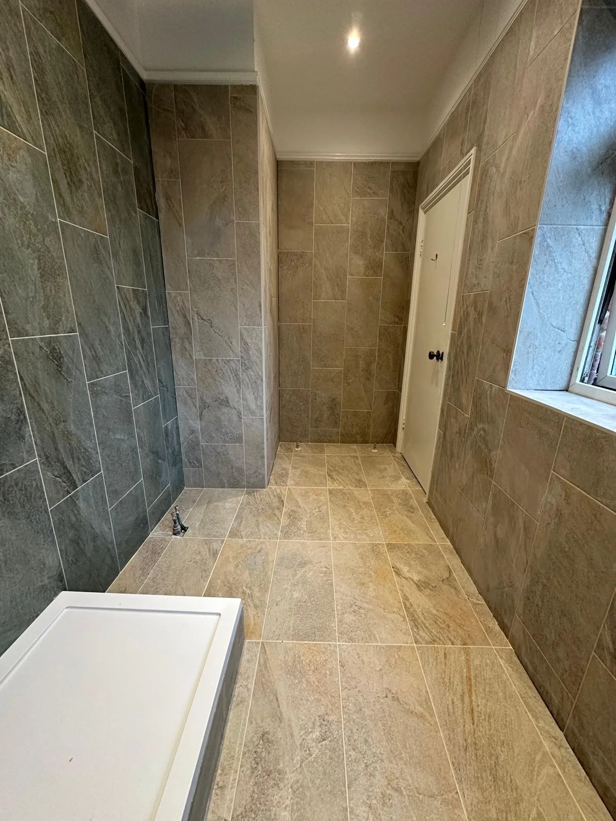 Empty shower area with beige and gray stone tiles on the walls and floor, a small rectangular shower base in the bottom left corner, a small window on the right wall, and a white door at the back.