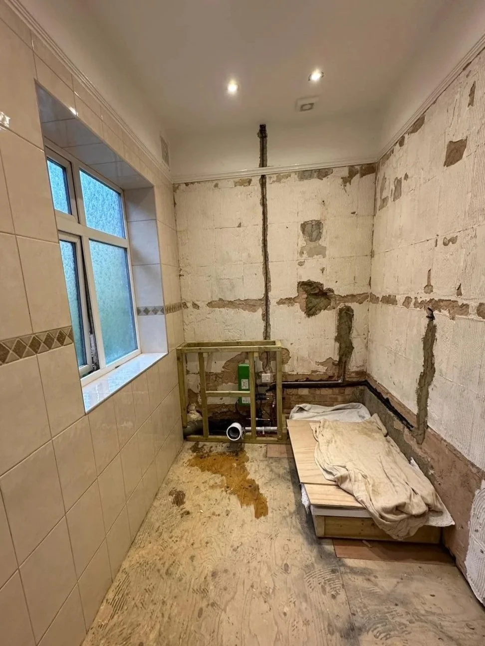 A room under renovation showing unfinished walls with exposed drywall, a partially tiled wall to the left, and plumbing fixtures in the corner, with construction materials on the floor.