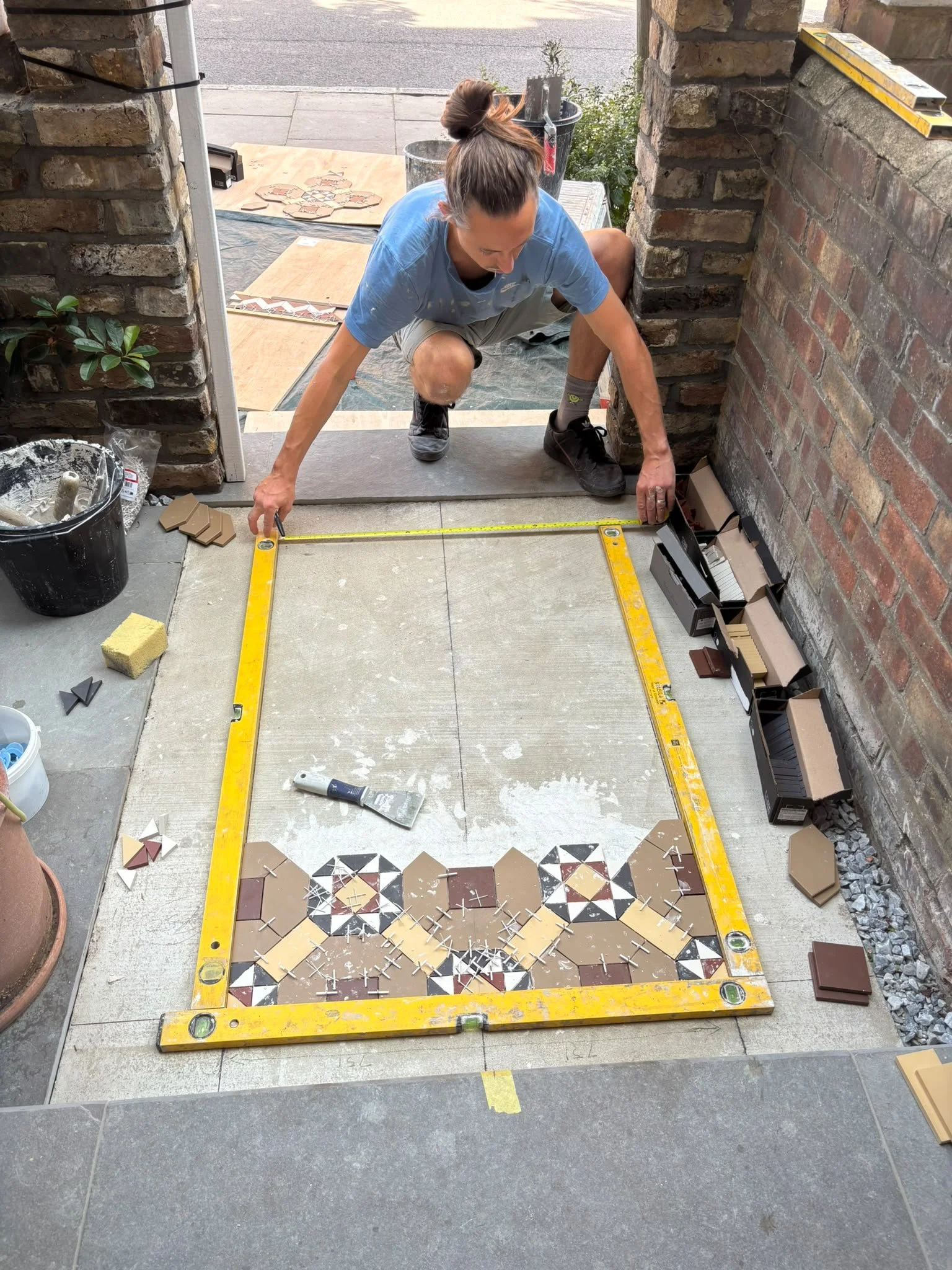 A woman is kneeling on the ground, measuring and installing decorative tiles on a section of floor outside a brick building. She is surrounded by tile tools, supplies, and a grid pattern of tiles on the floor.