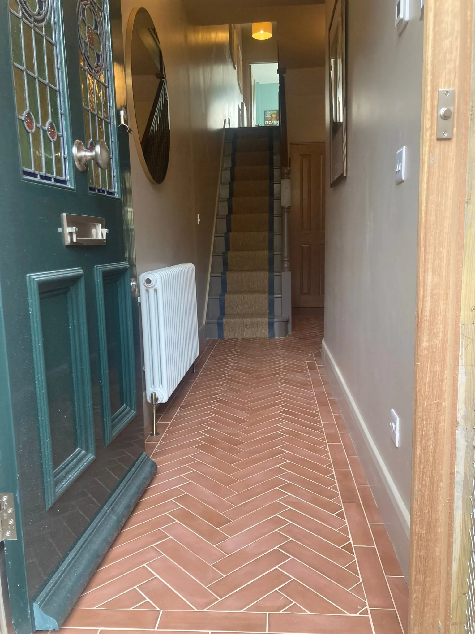 View of a hallway entrance with a green door, a heating radiator, a staircase with a beige and black runner, and brown herringbone tile flooring.
