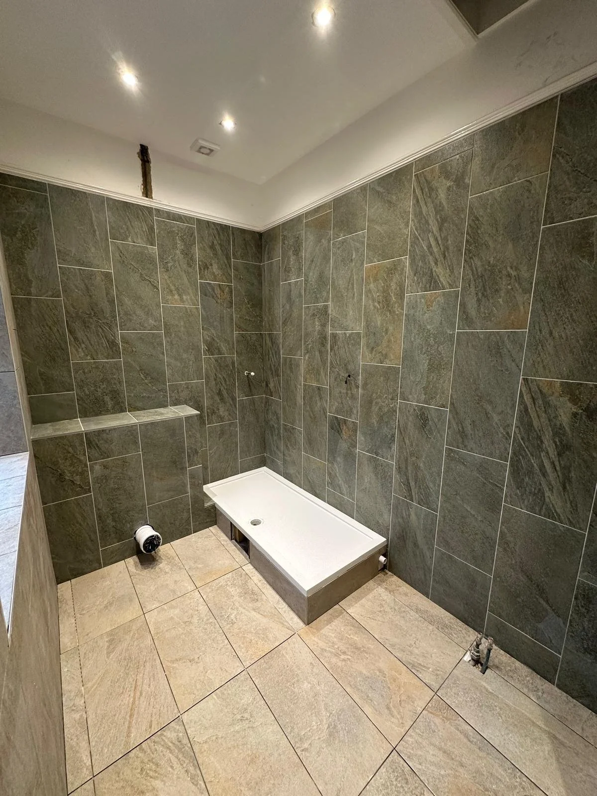 Bathroom under construction with dark green tiled walls and beige tiled floor, shower base installed, and unfinished fixtures.