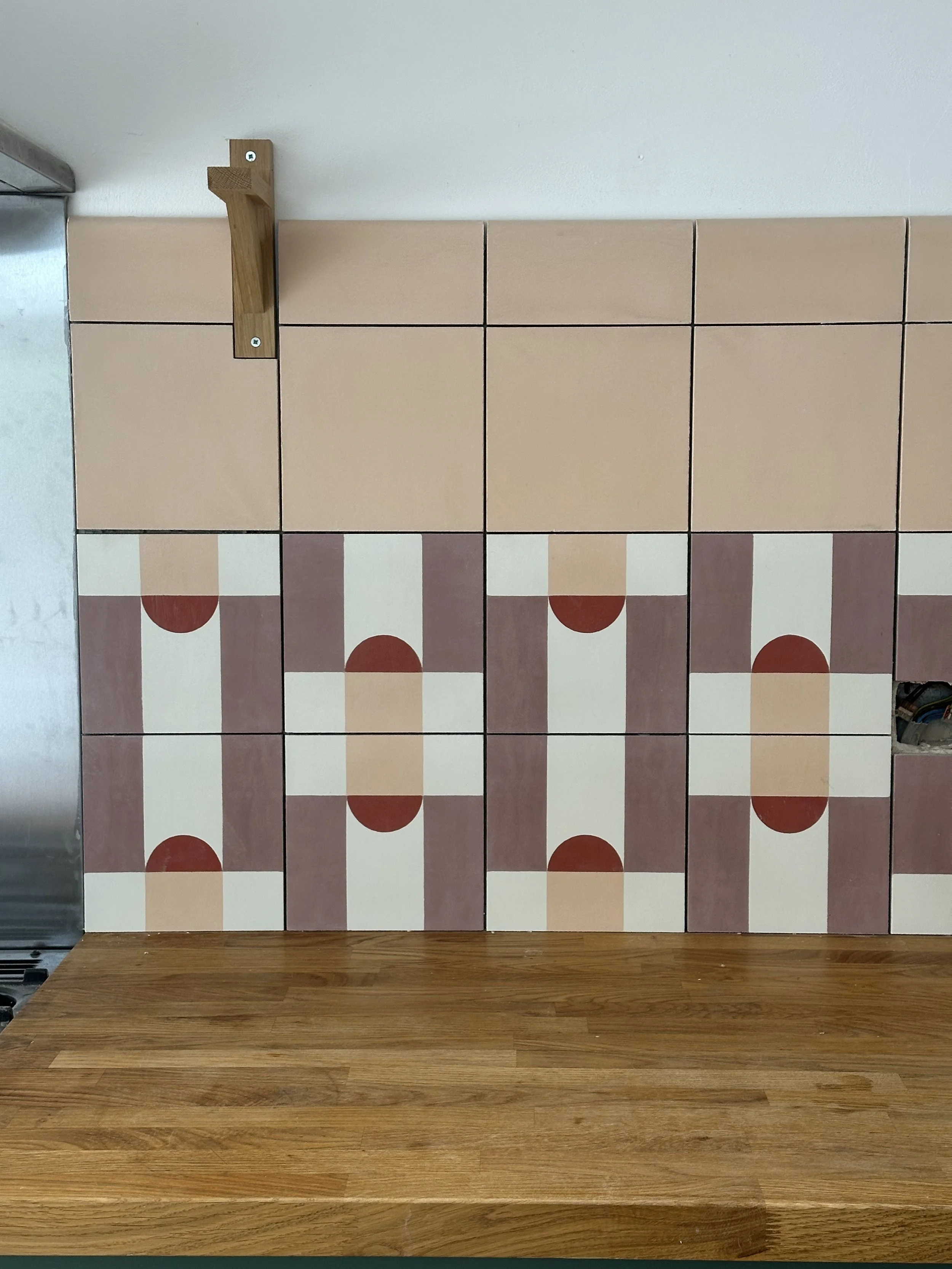 A wall with pink and patterned tiles, a wooden floor, and a metal appliance on the left.