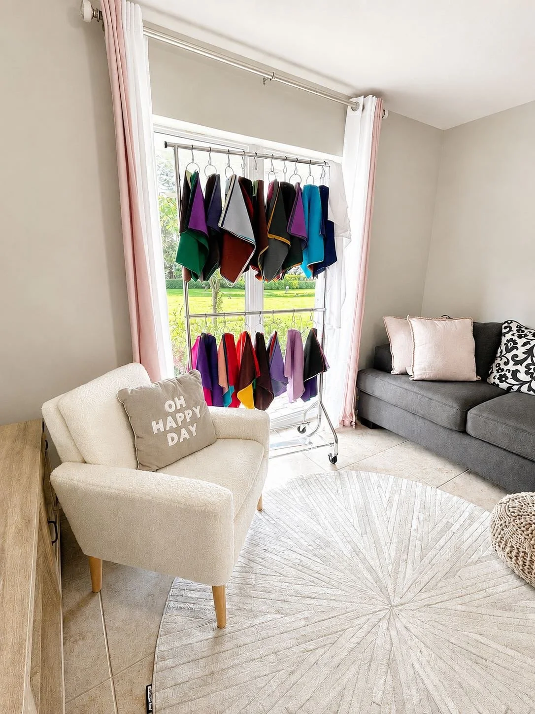 Room with a white armchair with a pillow that says 'OH HAPPY DAY' next to a gray sofa with pillows, and a clothing rack with multiple colorful cloth samples in front of a large window with pink curtains.
