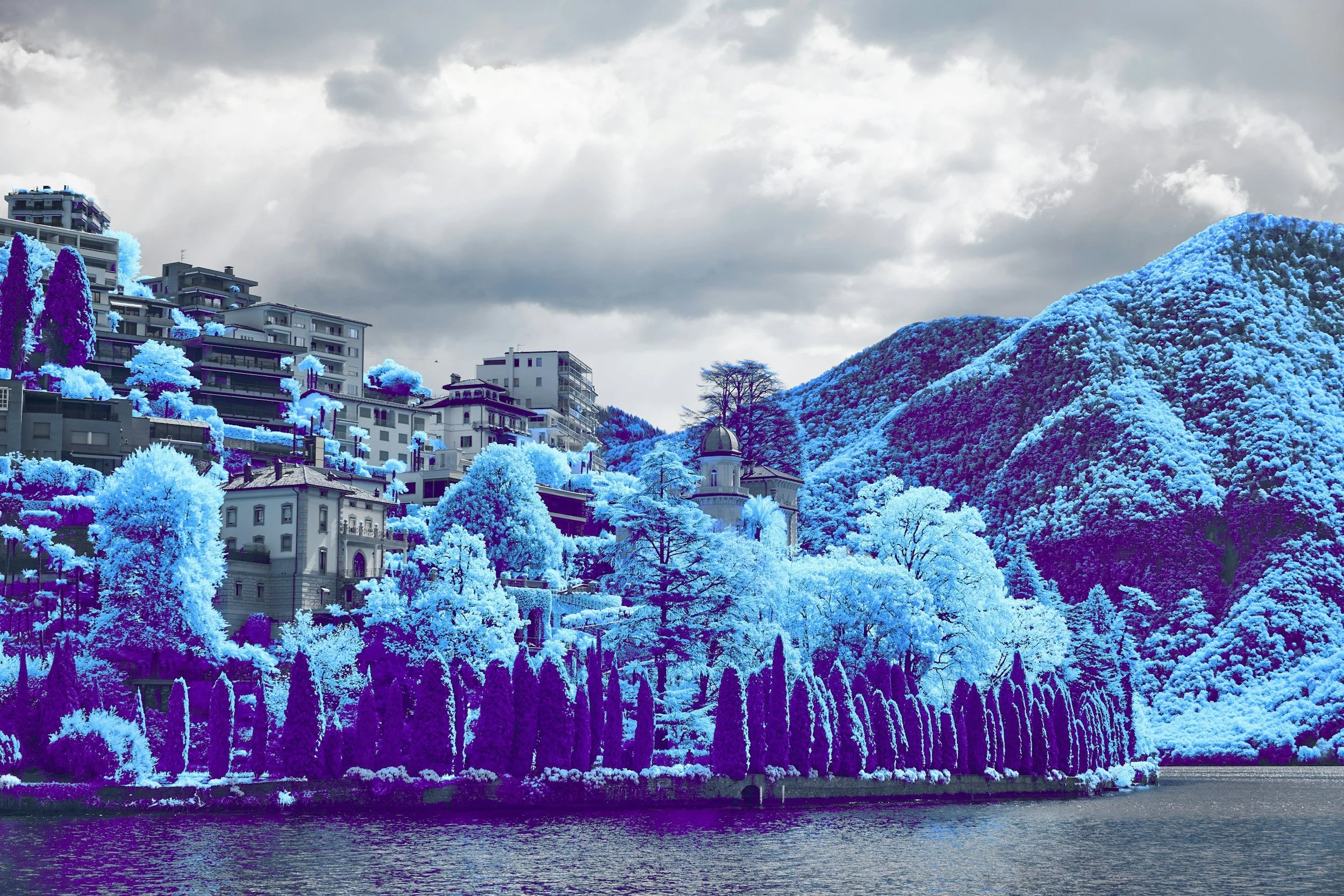 A cityscape with modern buildings on a hillside overlooking a body of water, with trees and landscape in color-inverted tones of blue and purple.