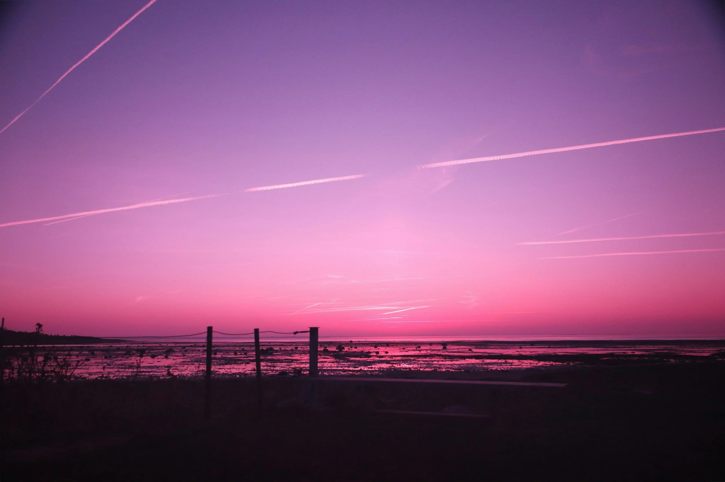A colorful sunset over a body of water with a fence in the foreground and contrails streaking across the purple and pink sky.
