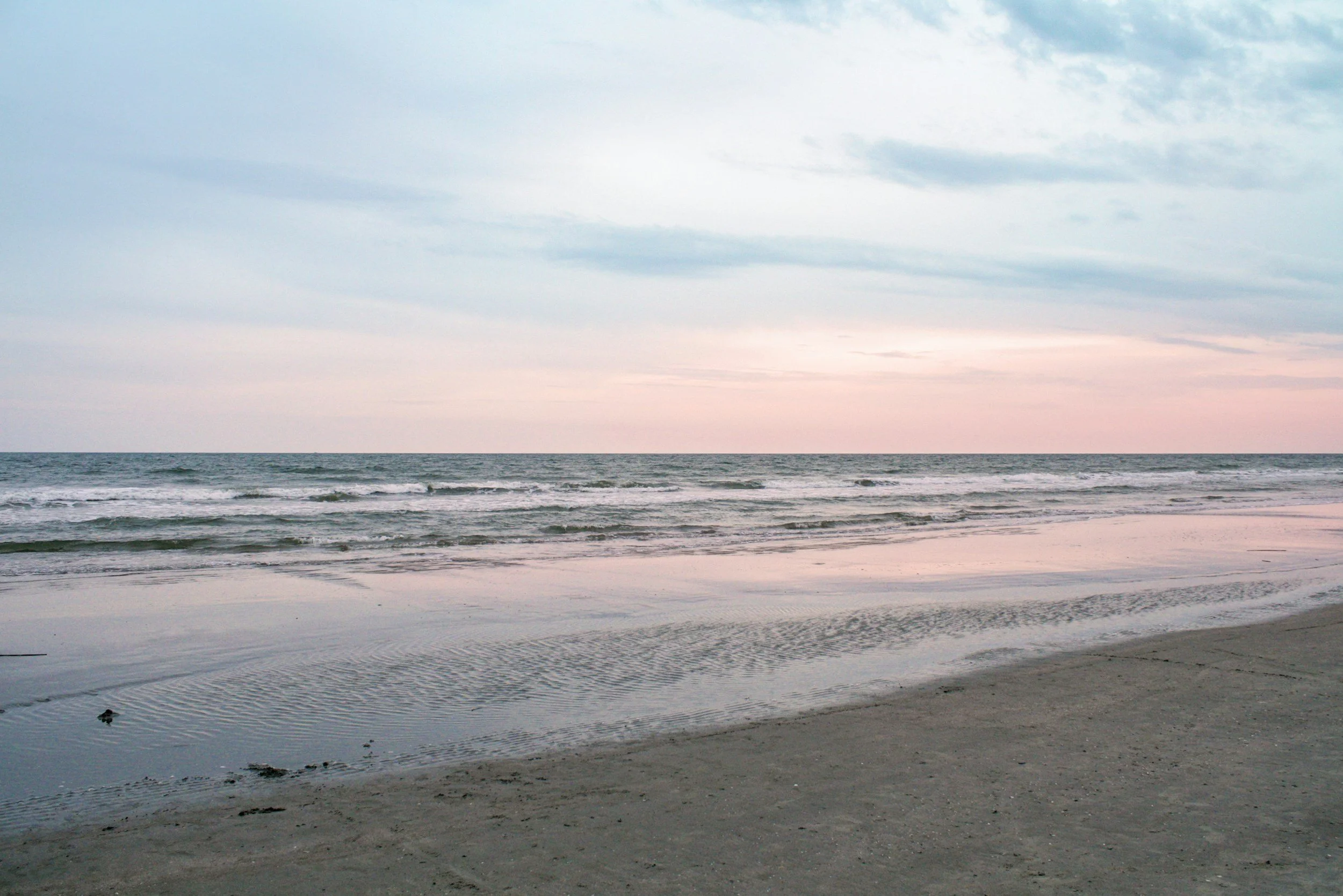 A beach with wet sand, gentle waves, and a pastel-colored sky with blue and pink hues.