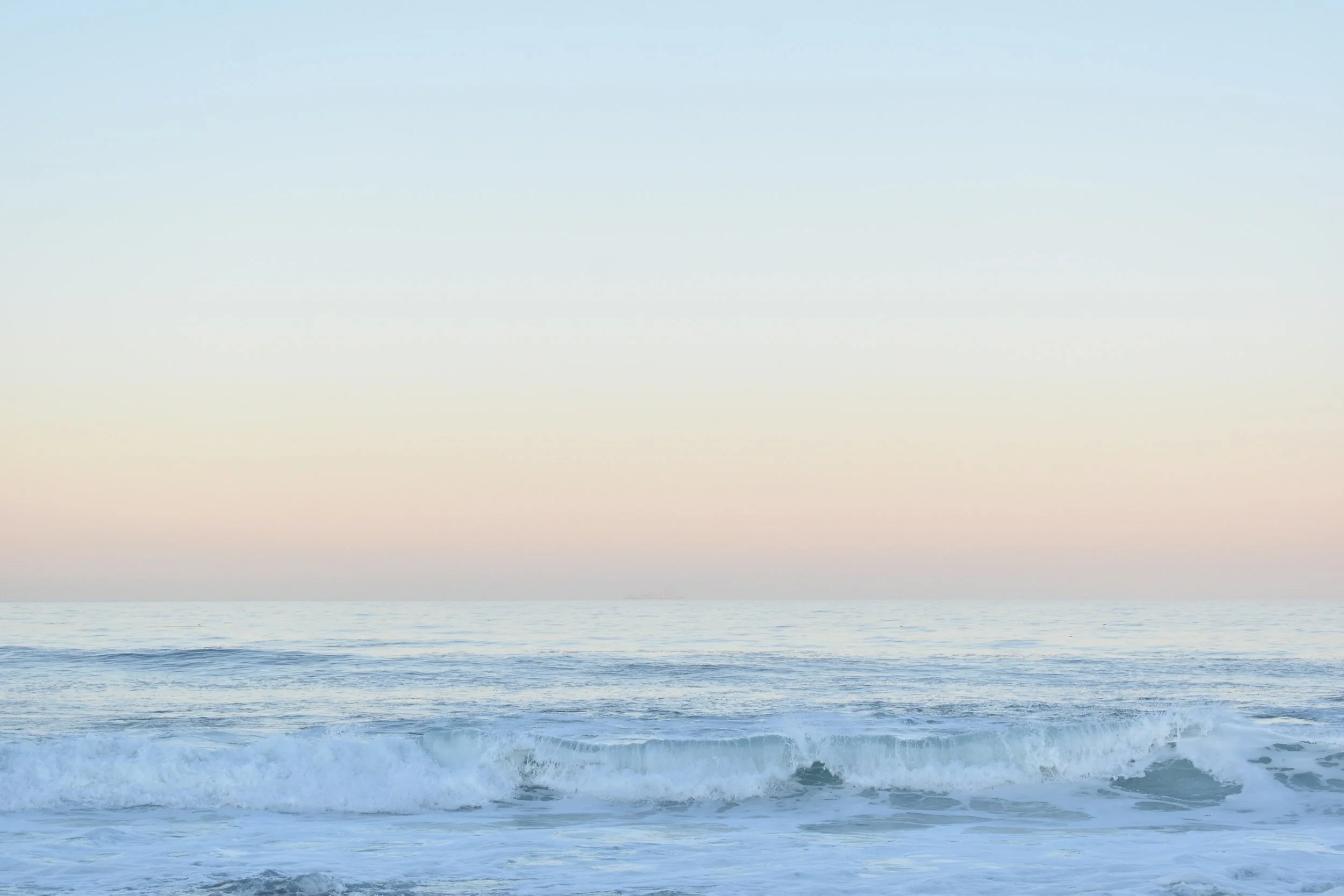 Calm ocean waves rolling onto the shore under a pastel-colored sky, possibly at dawn or dusk.