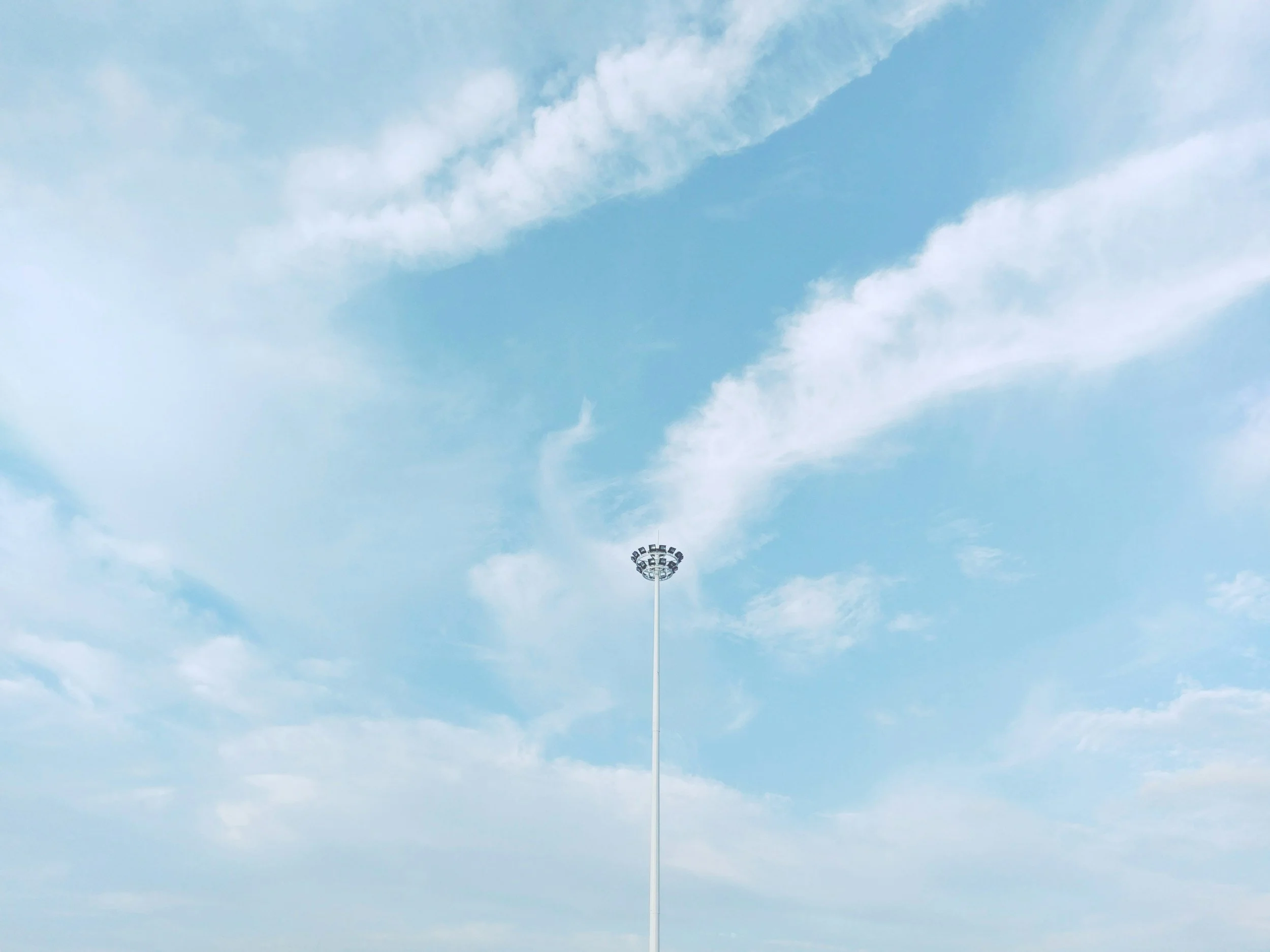 A clear blue sky with scattered white clouds and a tall light pole with multiple floodlights at the bottom center.