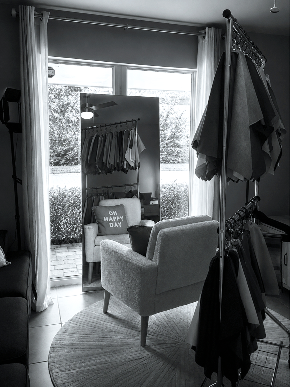 A black and white photo of a room with large window, curtains, armchair with pillow, standing mirror, and clothing rack with fabric samples.