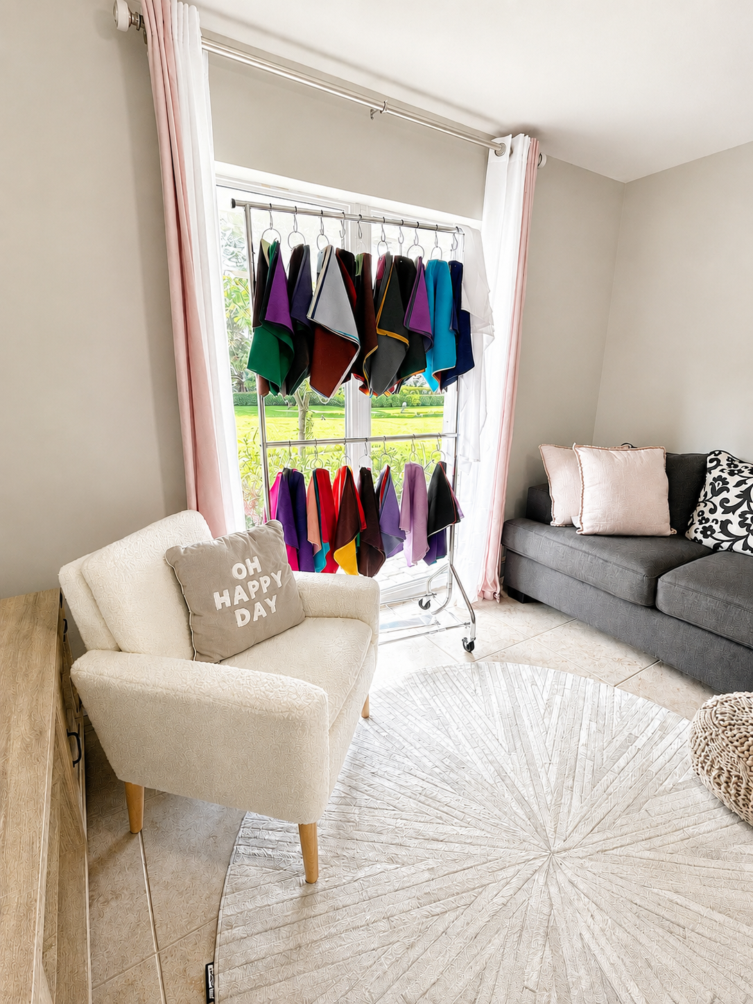 A living room with a white armchair and a gray sofa, a round beige rug, a blanket ladder with folded colorful towels, a window with pink curtains showing a green outdoor view, and throw pillows on the sofa.