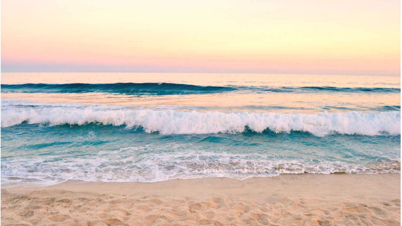 A sandy beach with gentle ocean waves crashing onto the shore during a pastel-colored sunset or sunrise.