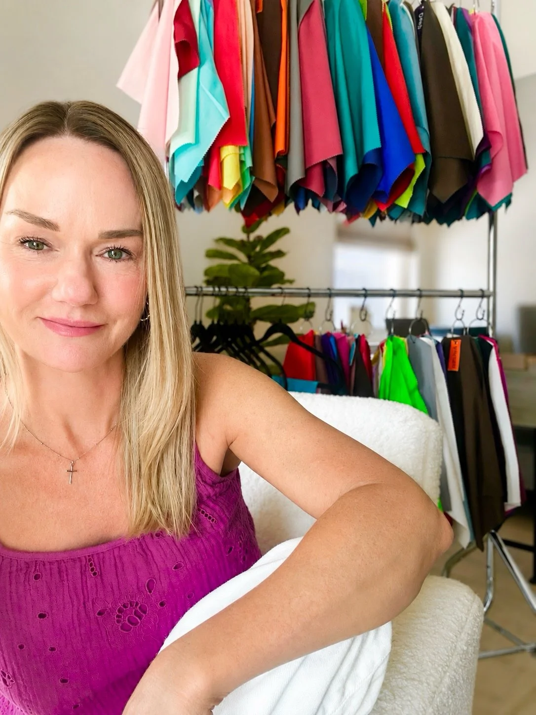 A woman with blonde hair sitting on a white chair, wearing a pink sleeveless top, and smiling at the camera. Behind her, there are colorful fabric swatches hanging on racks.