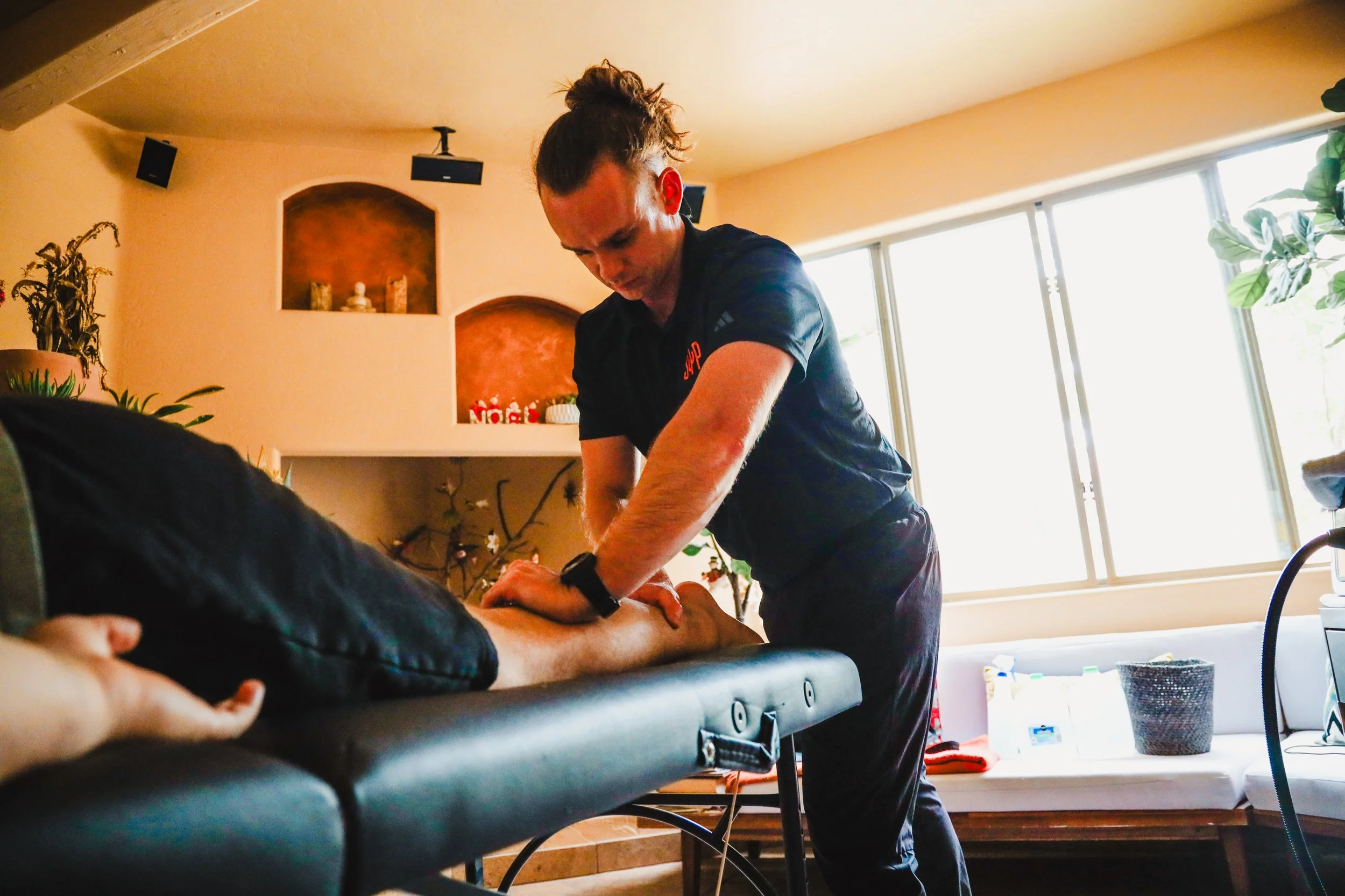 Person giving a massage to someone lying face down on a massage table in a brightly lit room with large windows and houseplants.
