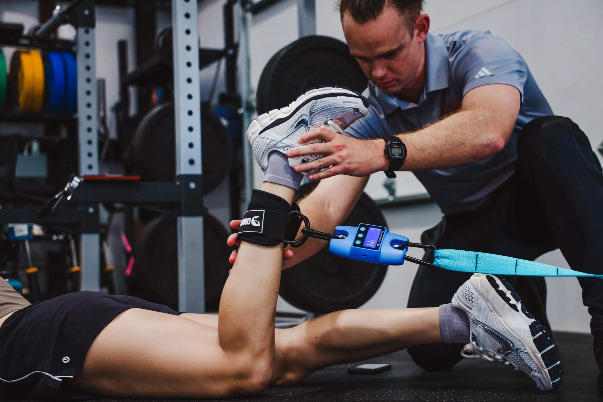 A fitness trainer helps a person during a stretching exercise in a gym. The trainer, wearing a light blue shirt and black shorts, holds the person's foot while they lie on the floor. The gym has exercise equipment and weights in the background.