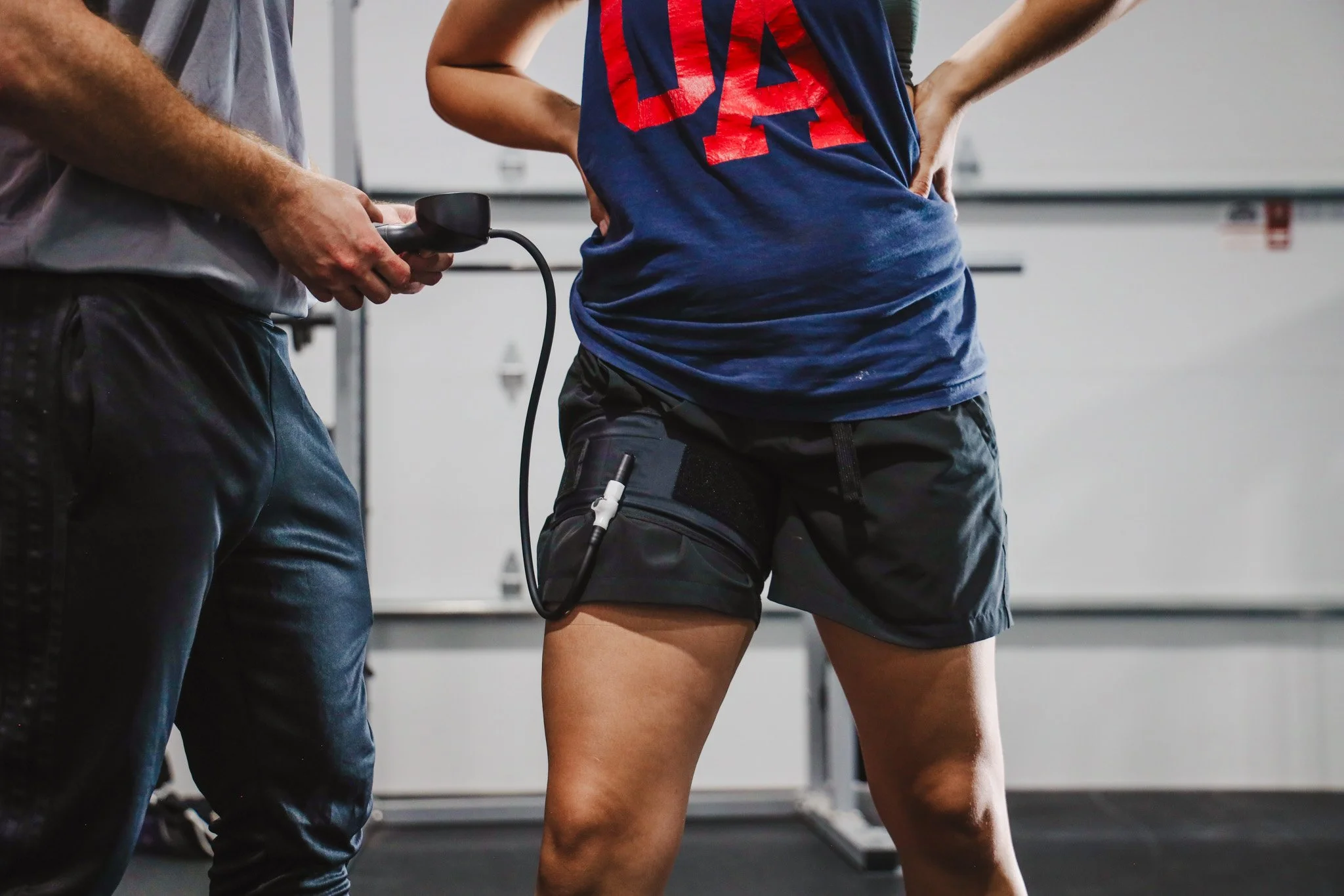 A person with muscular arms and a blue and red shirt standing with hands on hips next to another person holding a device, in a gym or training environment.