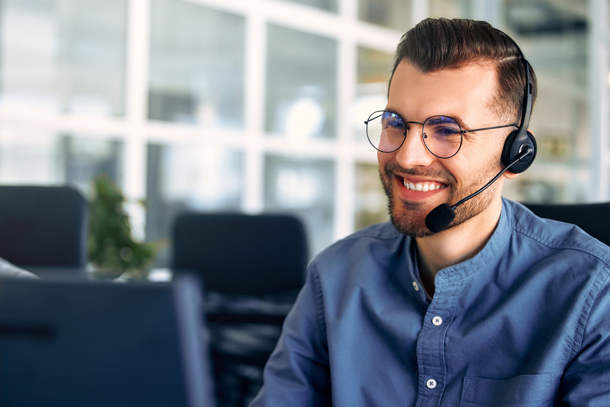 A young man wearing glasses and a headset with a microphone, smiling while working at his desk in an office.