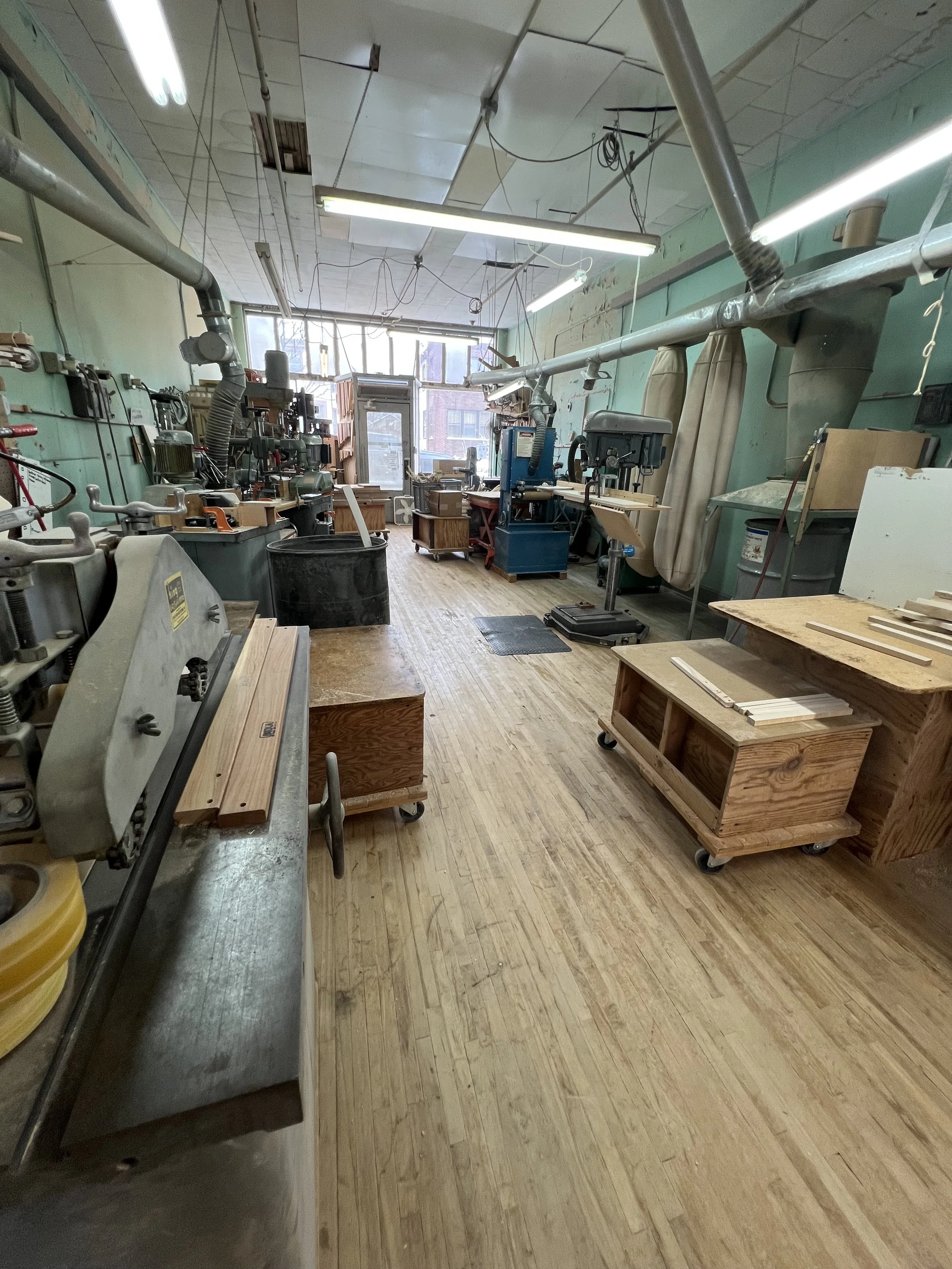 A woodworking or metalworking workshop with various machines and tools. There are wooden workbenches, a blue machine, and large pipes running along the ceiling. The room has large windows at the far end, letting in natural light, and the floor is made of wood.
