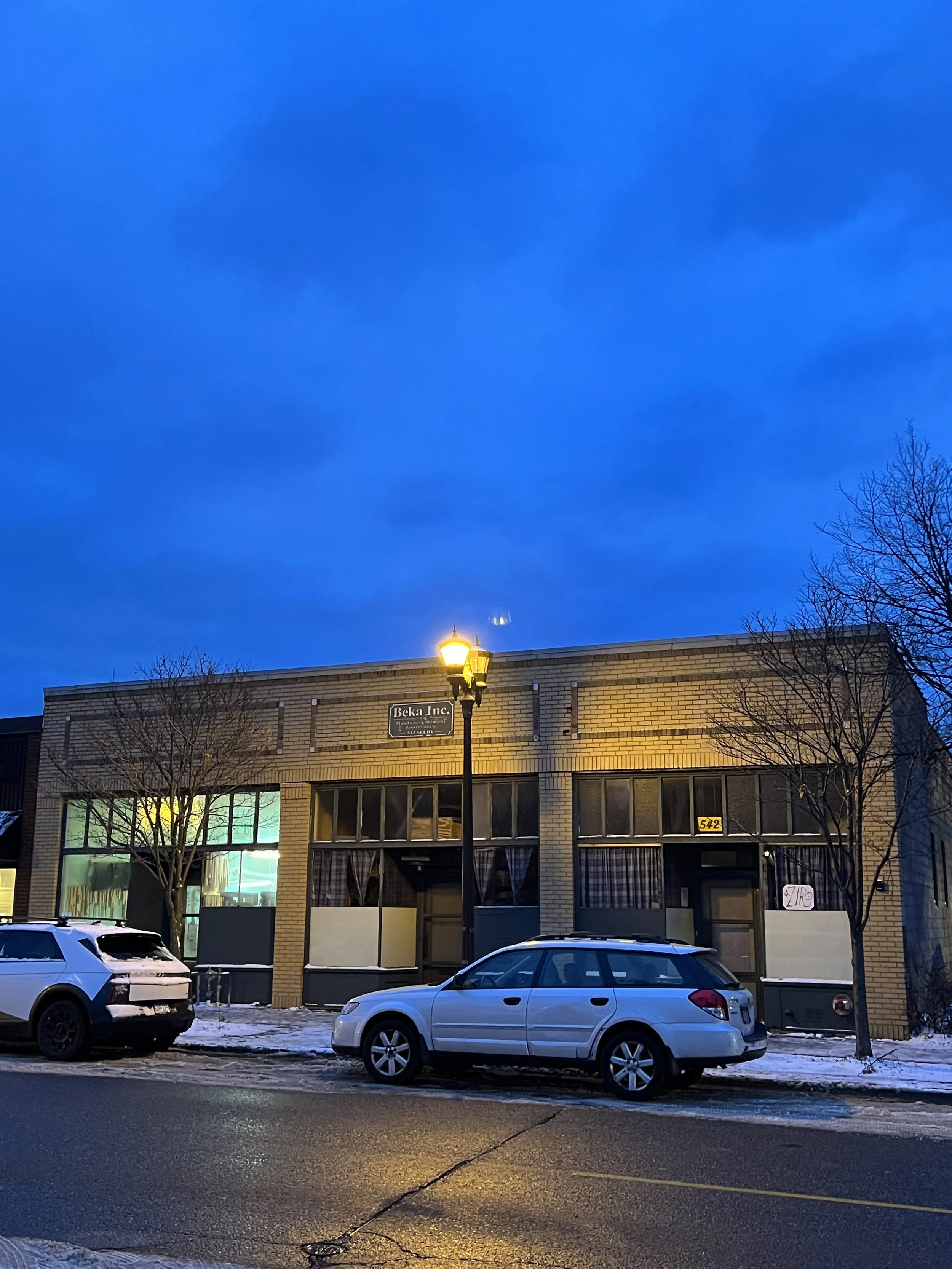 A street view at dusk showing a brick building with large windows and a sign that reads 'Beka Inc.' There are several parked cars in front of the building and a streetlamp illuminating the scene. The sky is dark blue with some clouds, and the trees are leafless, indicating winter.