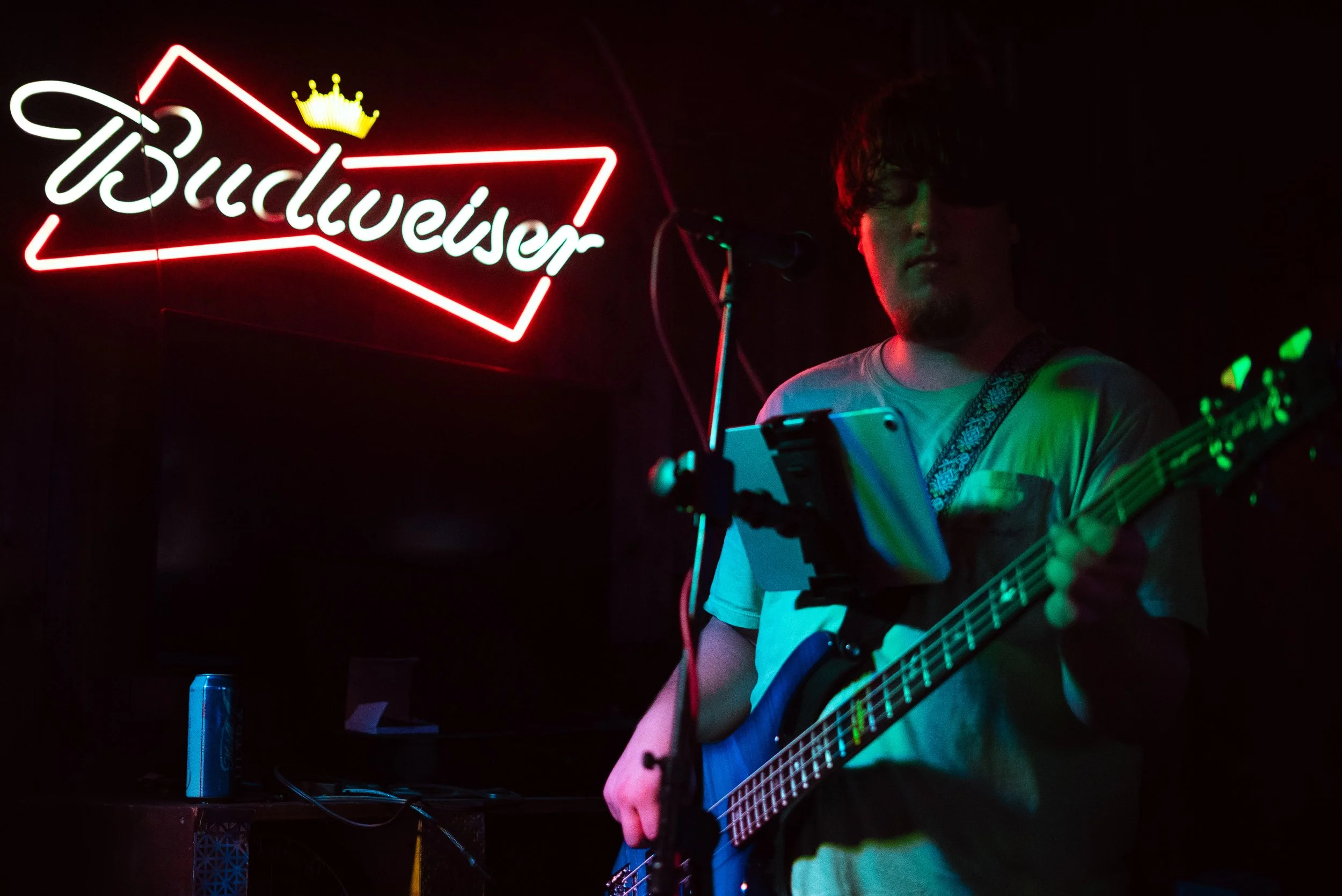 A person with curly hair and glasses playing an acoustic guitar on stage, illuminated by colorful stage lights, with a neon Budweiser sign in the background.