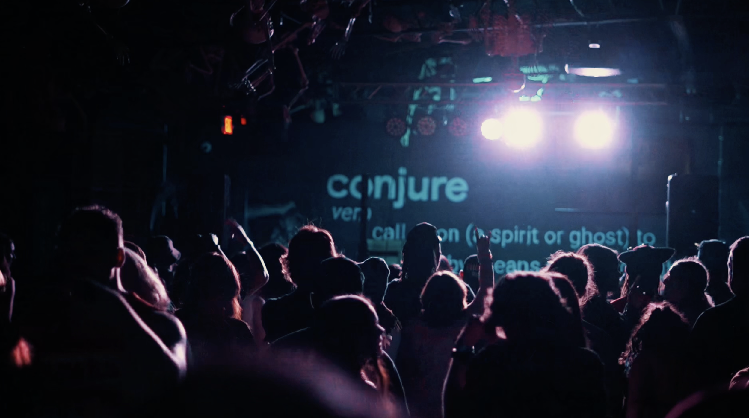 Crowd of people at a nighttime event with stage lights and screen displaying text, including the word 'conjure'.