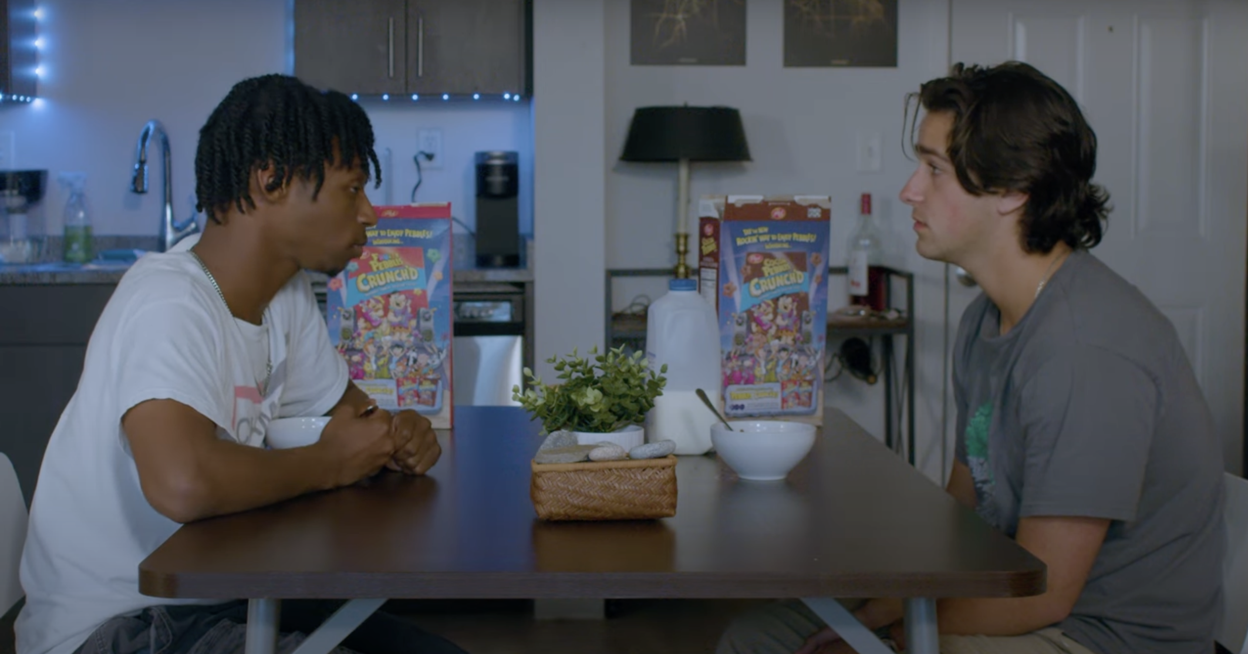 Two young men sitting across from each other at a kitchen dining table, engaging in conversation. The table has a small plant, a basket, and cereal boxes in the background.