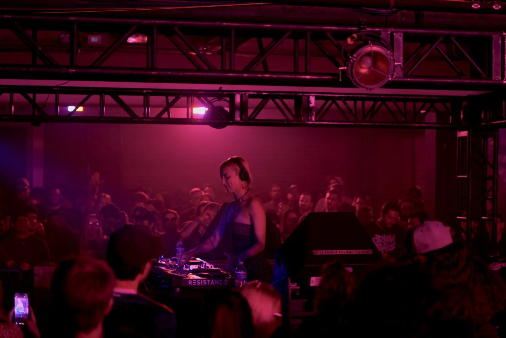 A female DJ performing on stage with pink lighting, surrounded by a crowd of people at a nightclub or concert.