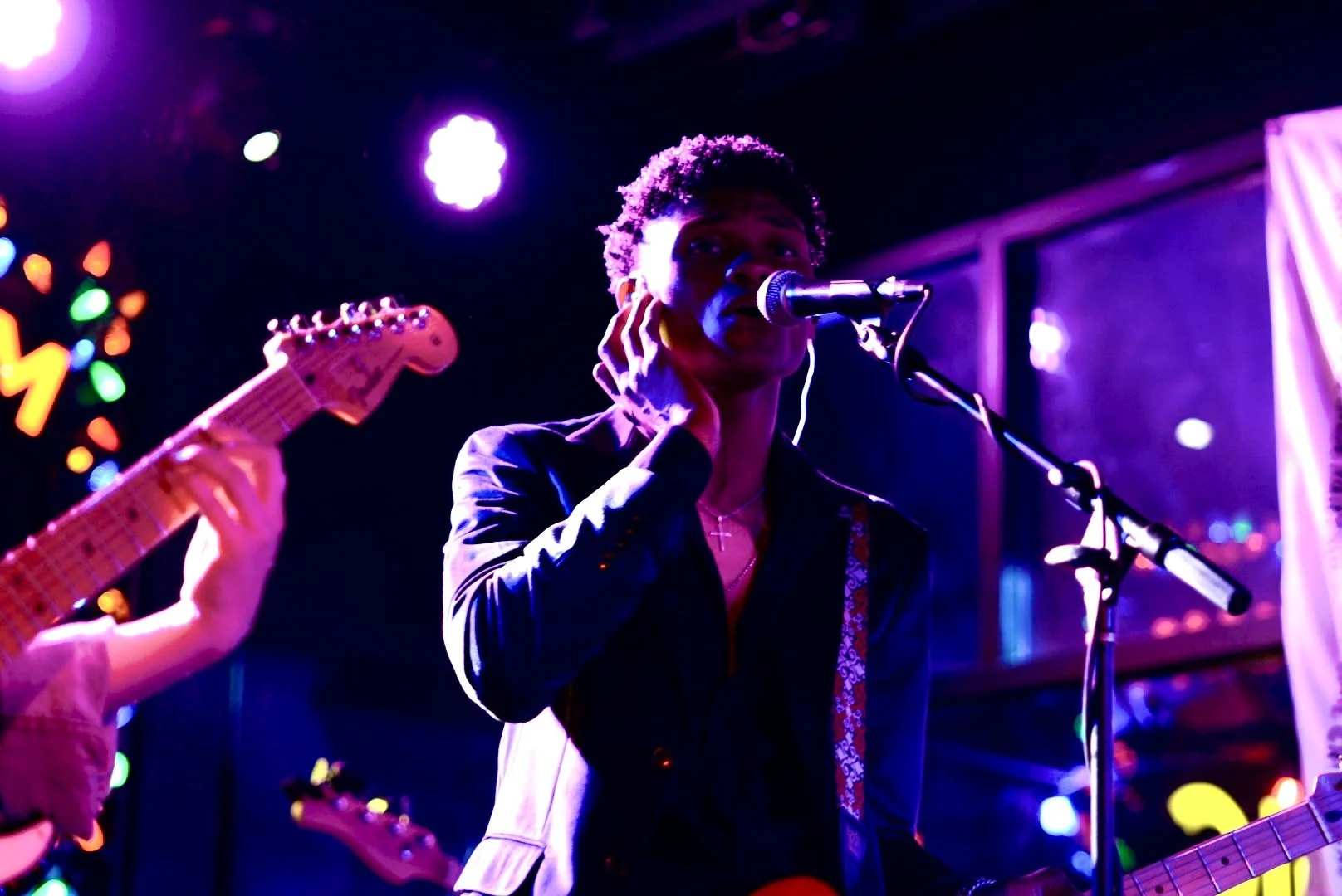 A male musician singing into a microphone while playing guitar on stage, with colorful lights and a dark background.