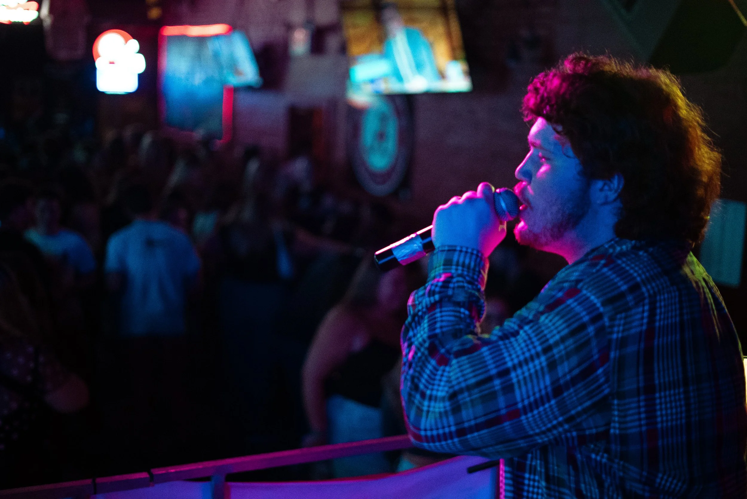 A person with curly hair, wearing a checkered shirt, holding a microphone, appears to be performing or speaking at a nighttime event with a crowd and neon lights in the background.