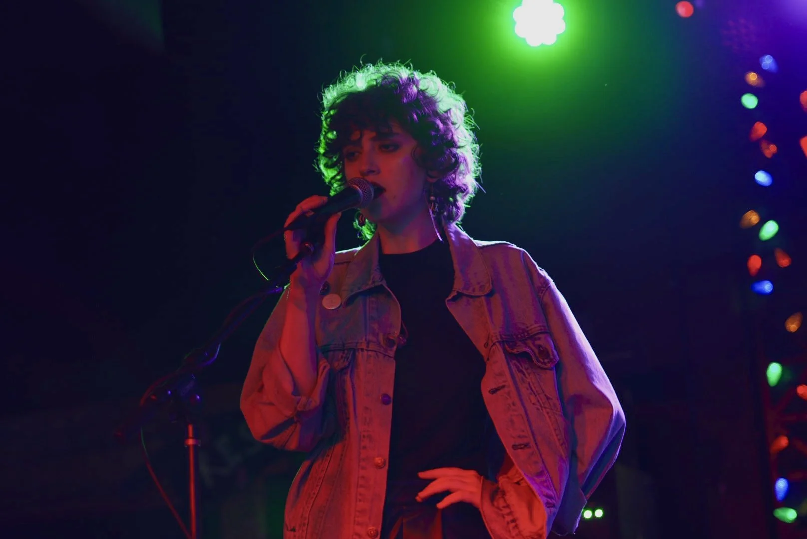 A woman with curly hair wearing a denim jacket singing into a microphone on stage with colorful lights in the background.
