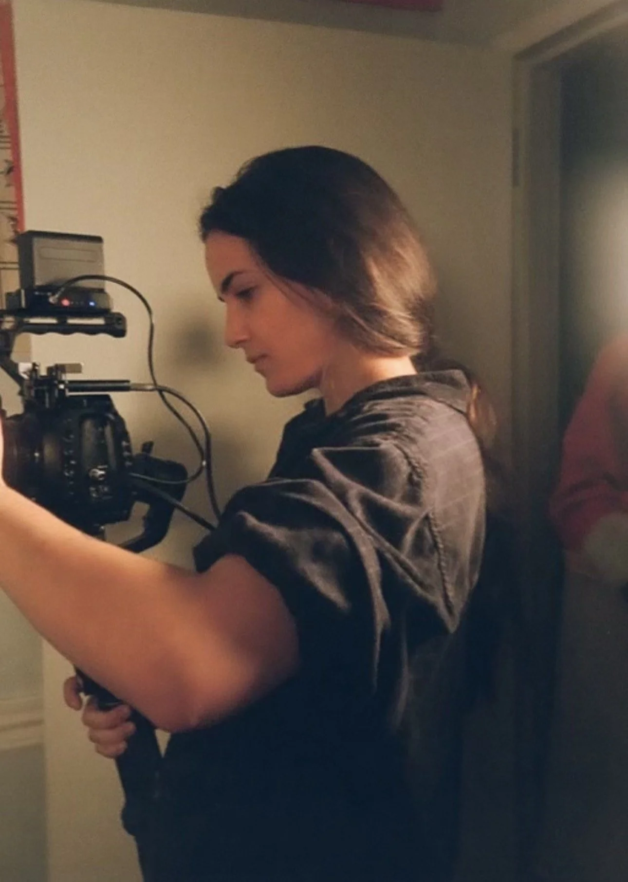 A woman with shoulder-length dark hair operating a professional camera on a tripod, wearing a dark shirt with rolled-up sleeves, in a room with beige walls.