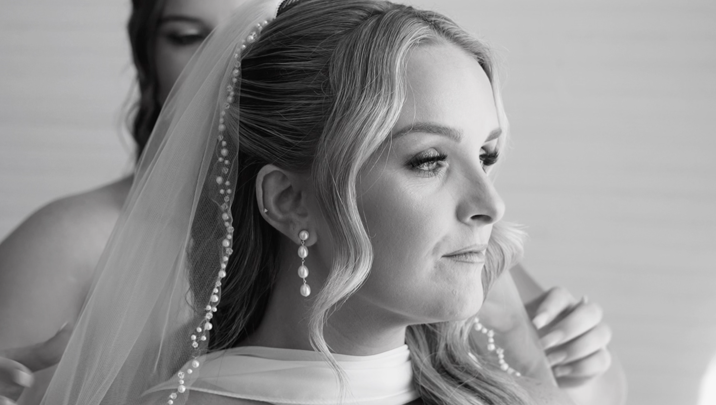 Black and white close-up of a bride wearing a veil with pearls, earrings, and makeup, with another woman behind her adjusting the veil.