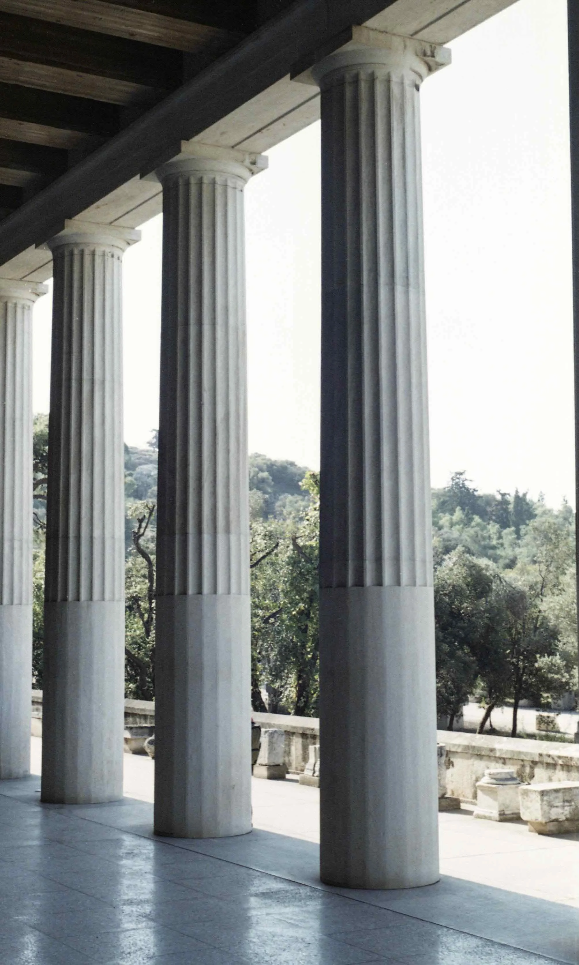 Four ancient Greek-style marble columns with fluted shafts supporting a roof, overlooking a landscape with trees and a clear sky.