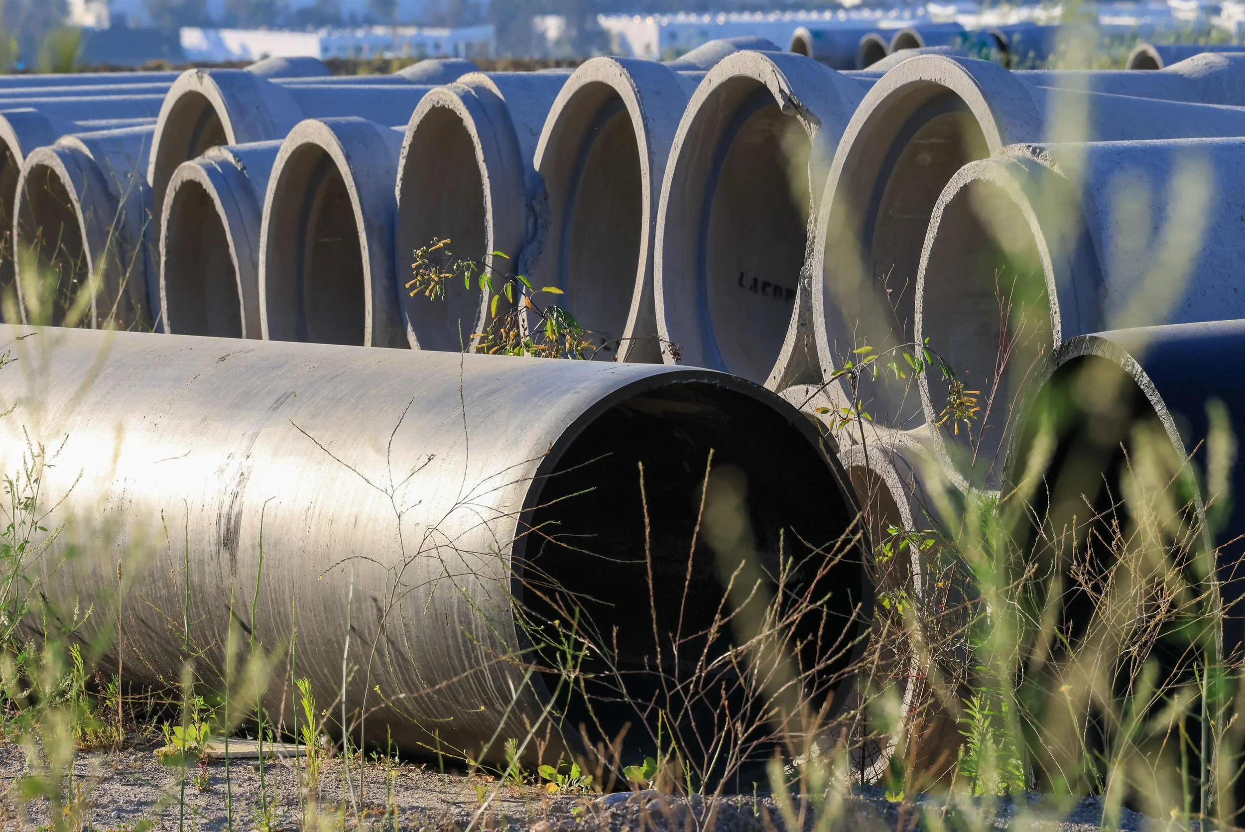 Large concrete pipes stacked outdoors with some grass and plants growing around them, under a clear sky.