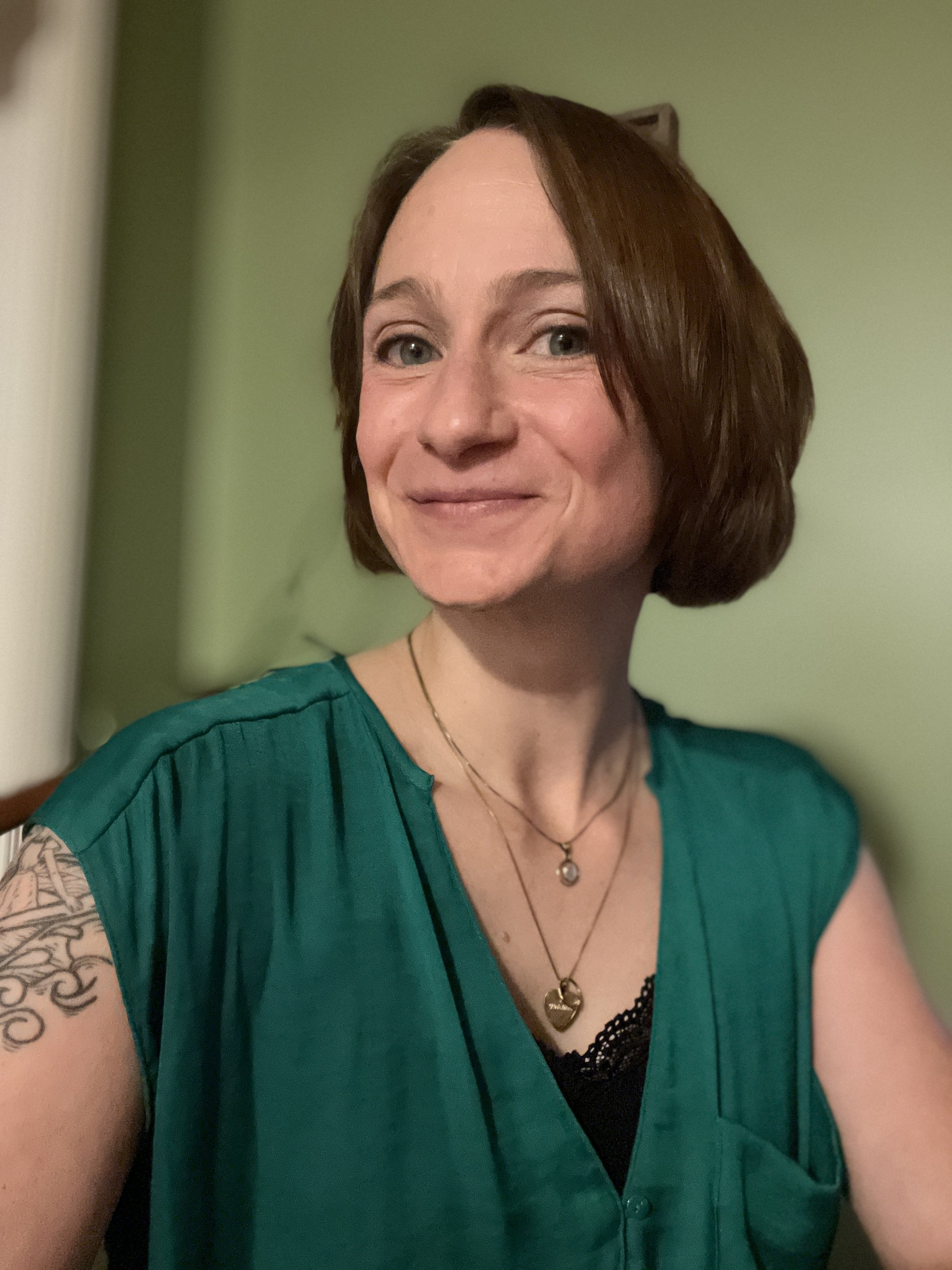 Close-up of a woman with short, brown hair, wearing a green top and layered necklaces, smiling softly, with a tattoo visible on her left arm against a green wall background.