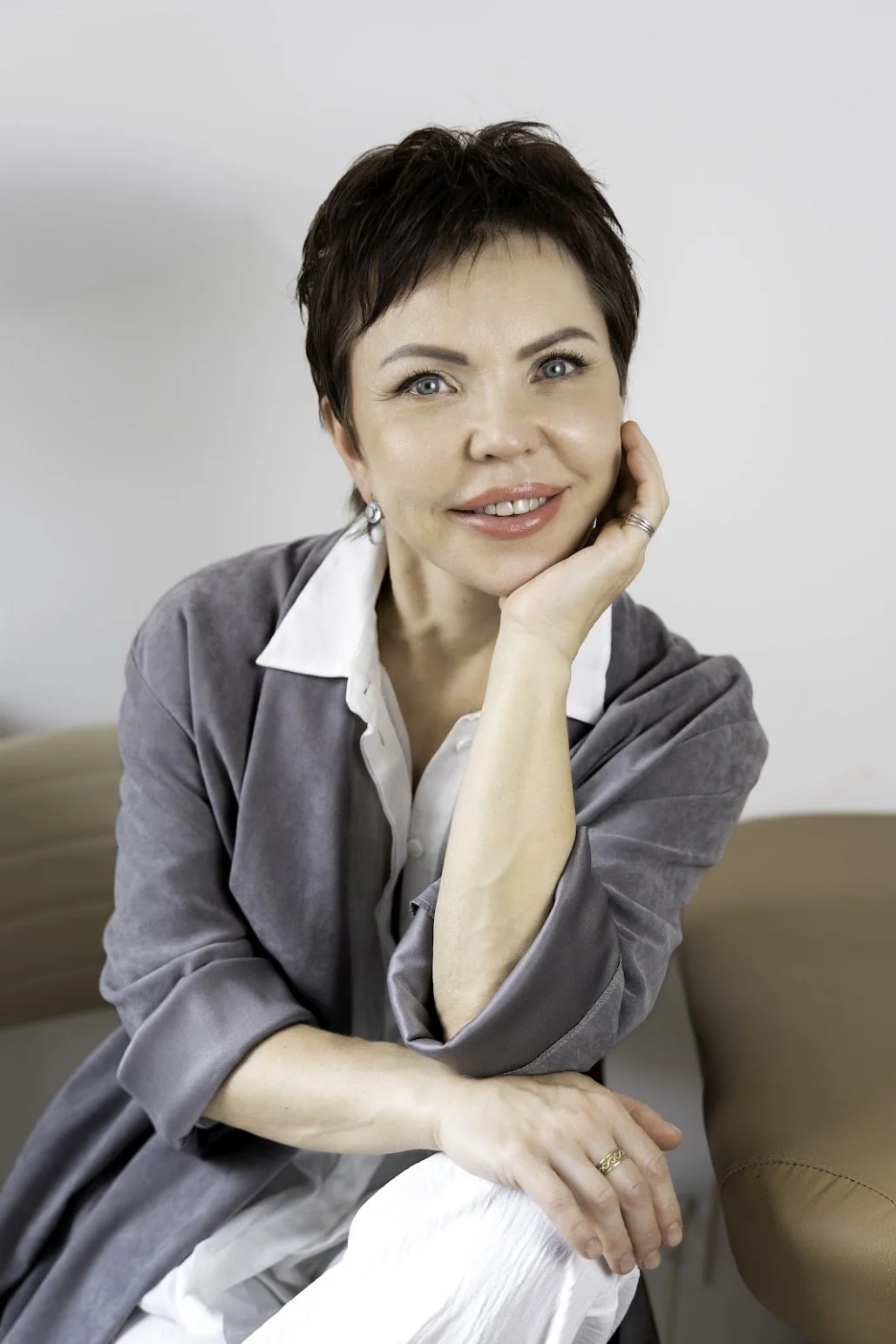 A woman with short dark hair and blue eyes, wearing a white shirt and grey cardigan, sitting on a beige couch, resting her face on her right hand, smiling gently at the camera.