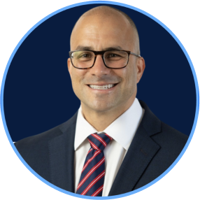 Portrait of a bald man wearing glasses, a dark suit, white shirt, and a red and blue striped tie, smiling against a dark blue background.