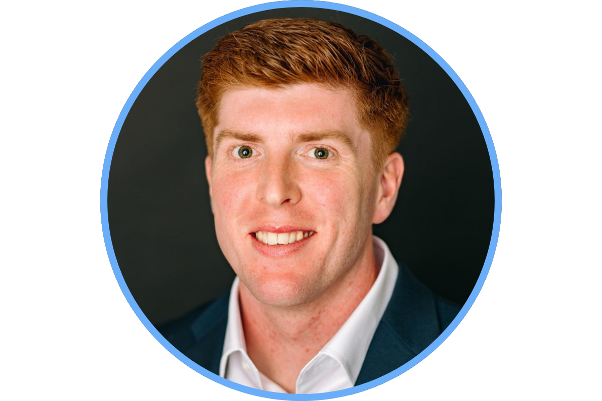 A headshot of a man with red hair, wearing a white shirt and dark suit jacket, smiling against a dark background.