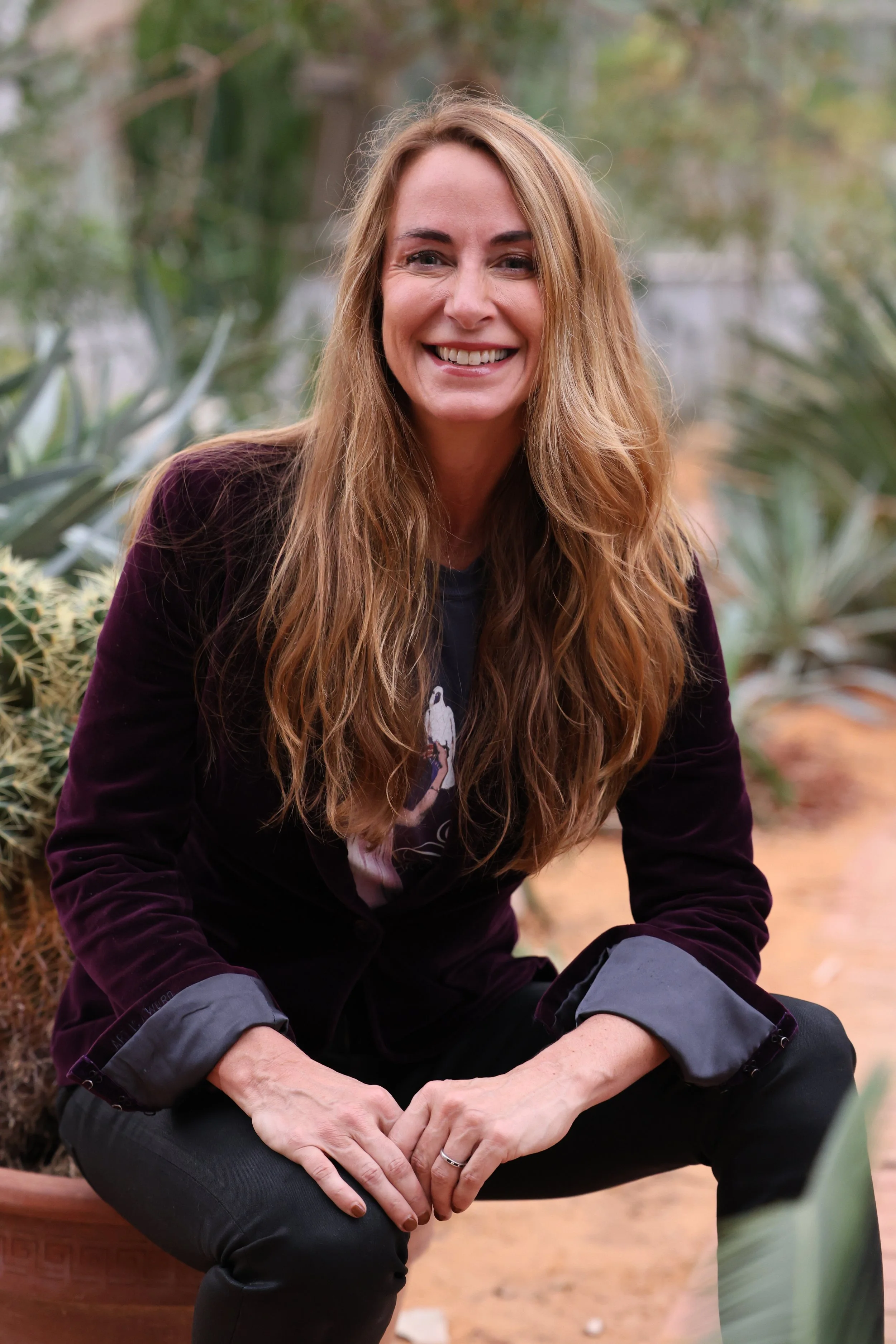 A smiling woman with long, wavy, red hair, wearing a dark jacket and black pants, squatting outdoors among desert plants.