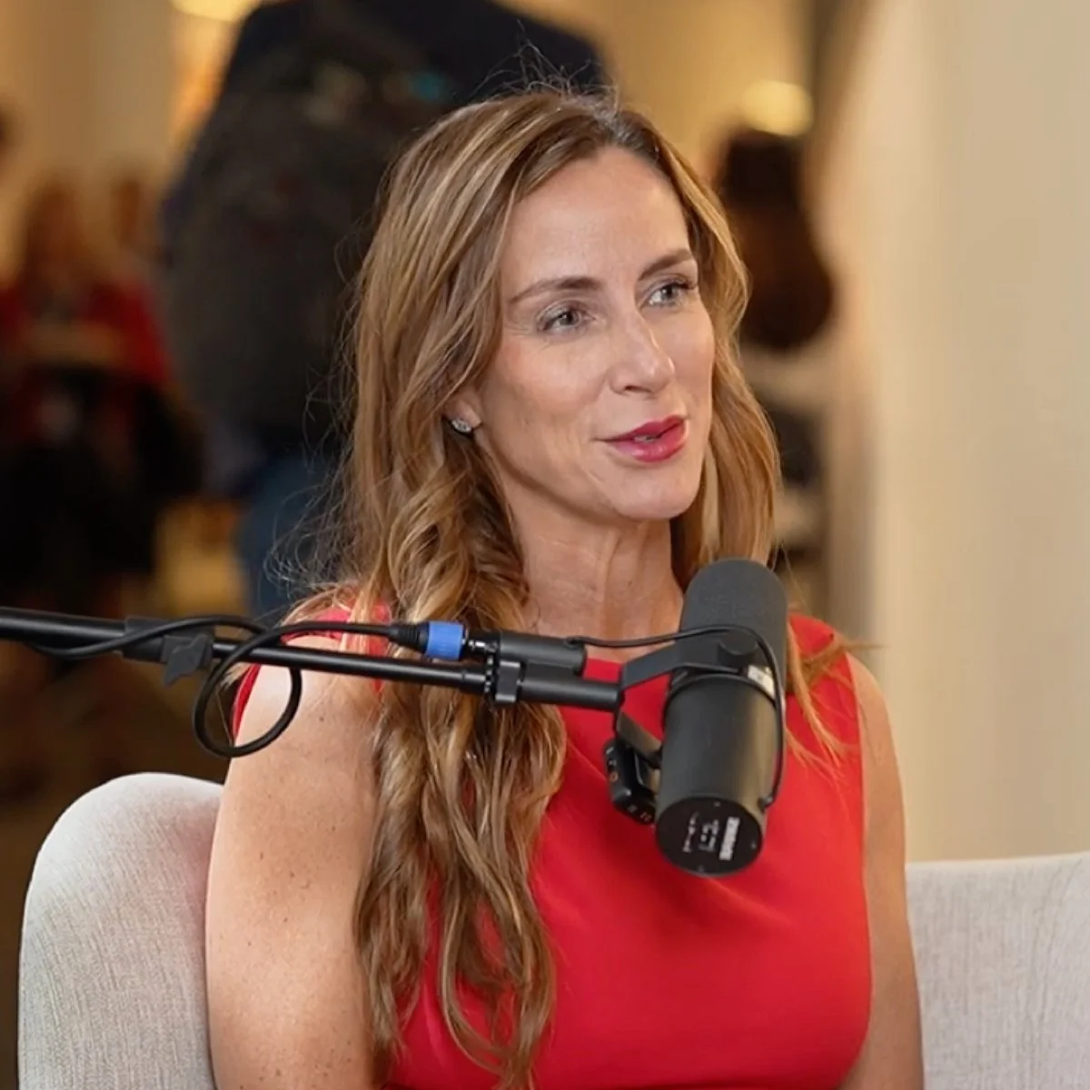 Woman with long wavy red hair speaking into a microphone during an interview or podcast, wearing a red sleeveless dress, seated on a light-colored chair, with a warm background and a blurred person in the background.