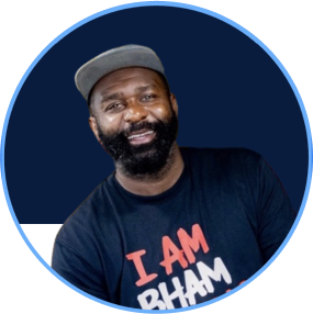 Portrait of a man with a beard wearing a gray cap and a black T-shirt that says 'I AM BHAM,' smiling against a blue background.