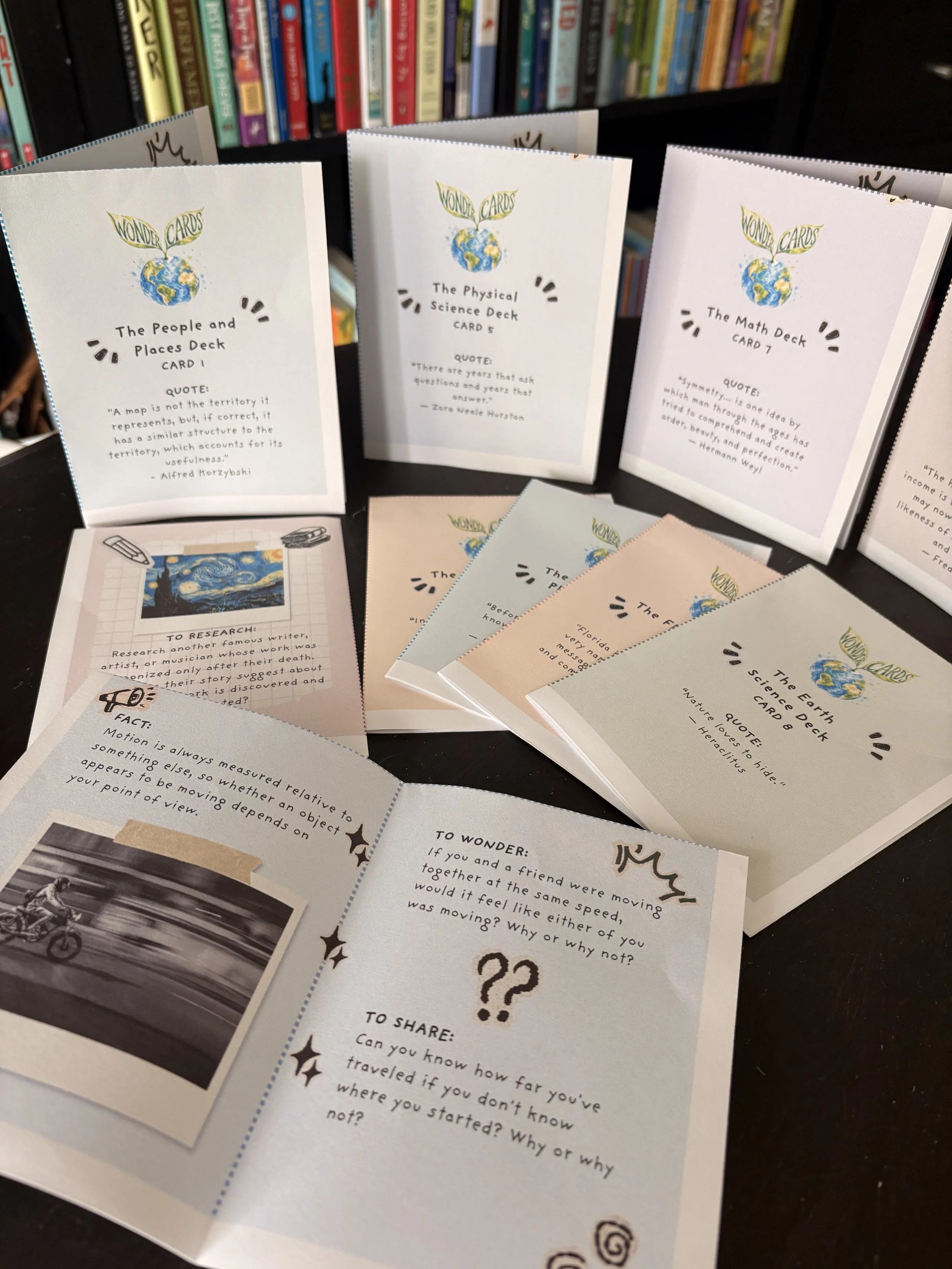 Multiple small pamphlets or cards with a 'Wonder Cards' logo featuring a globe with leaves, placed on a table with a bookshelf in the background. The cards contain questions and quotes related to science and curiosity.