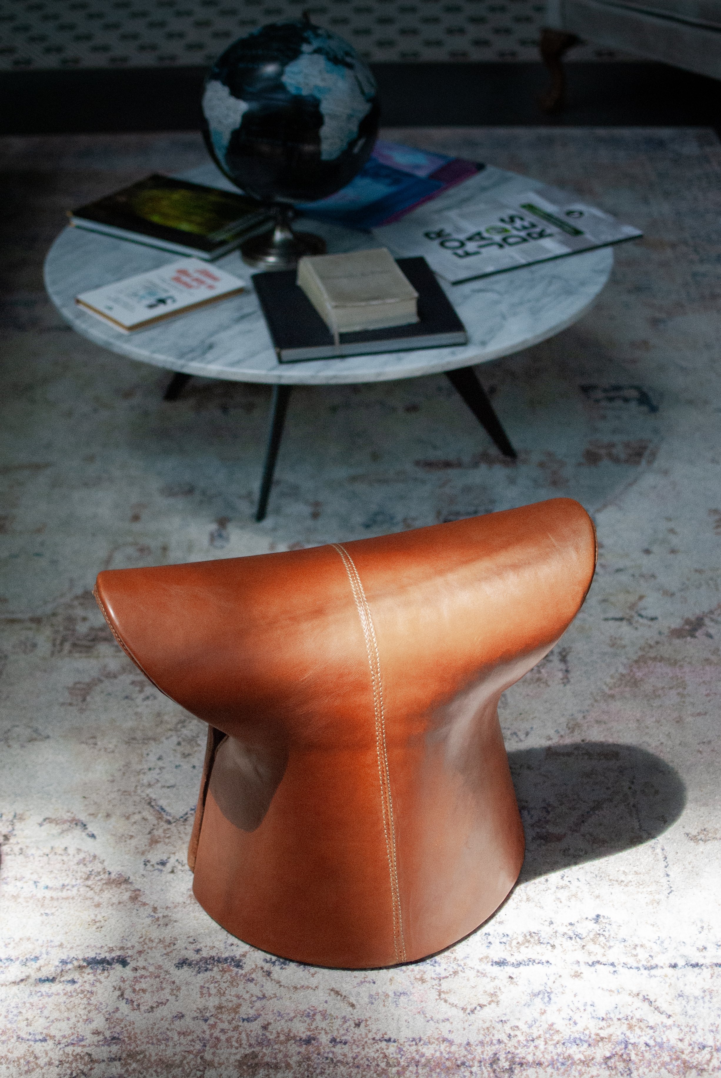 A round, modern lounge chair with a tan leather finish, placed on a textured, multicolored area rug. The chair has a curved back and seat.