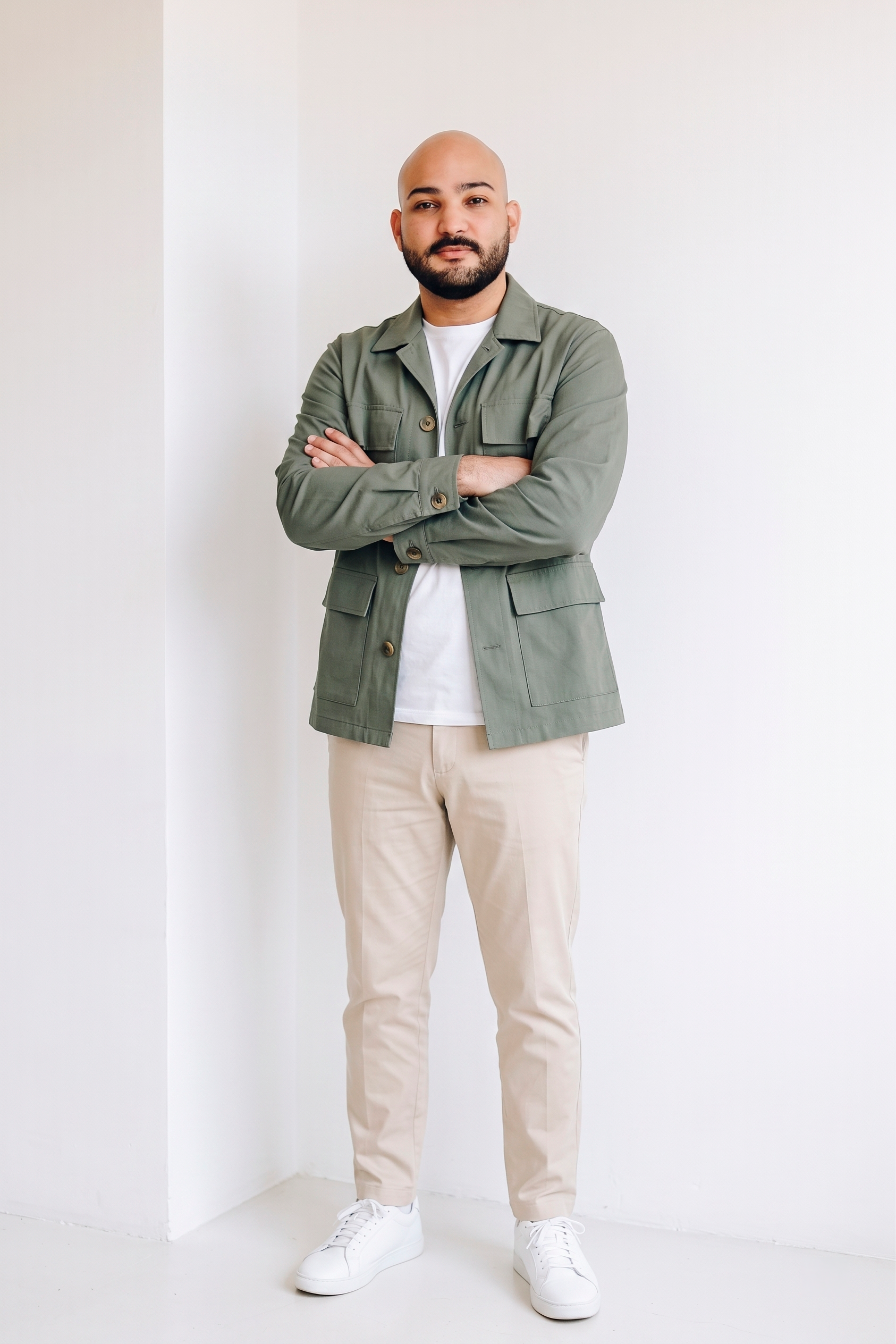 A man with a beard and bald head wearing a light green jacket, white T-shirt, beige pants, and white sneakers, standing with arms crossed against a plain white wall.