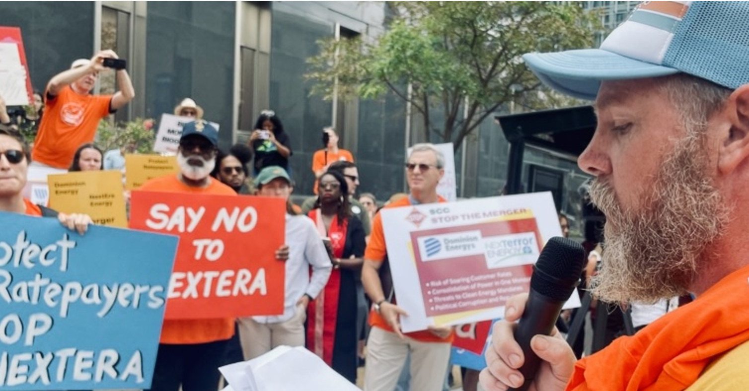man speaks at a rally with people holding signs opposing the nextera dominion merger