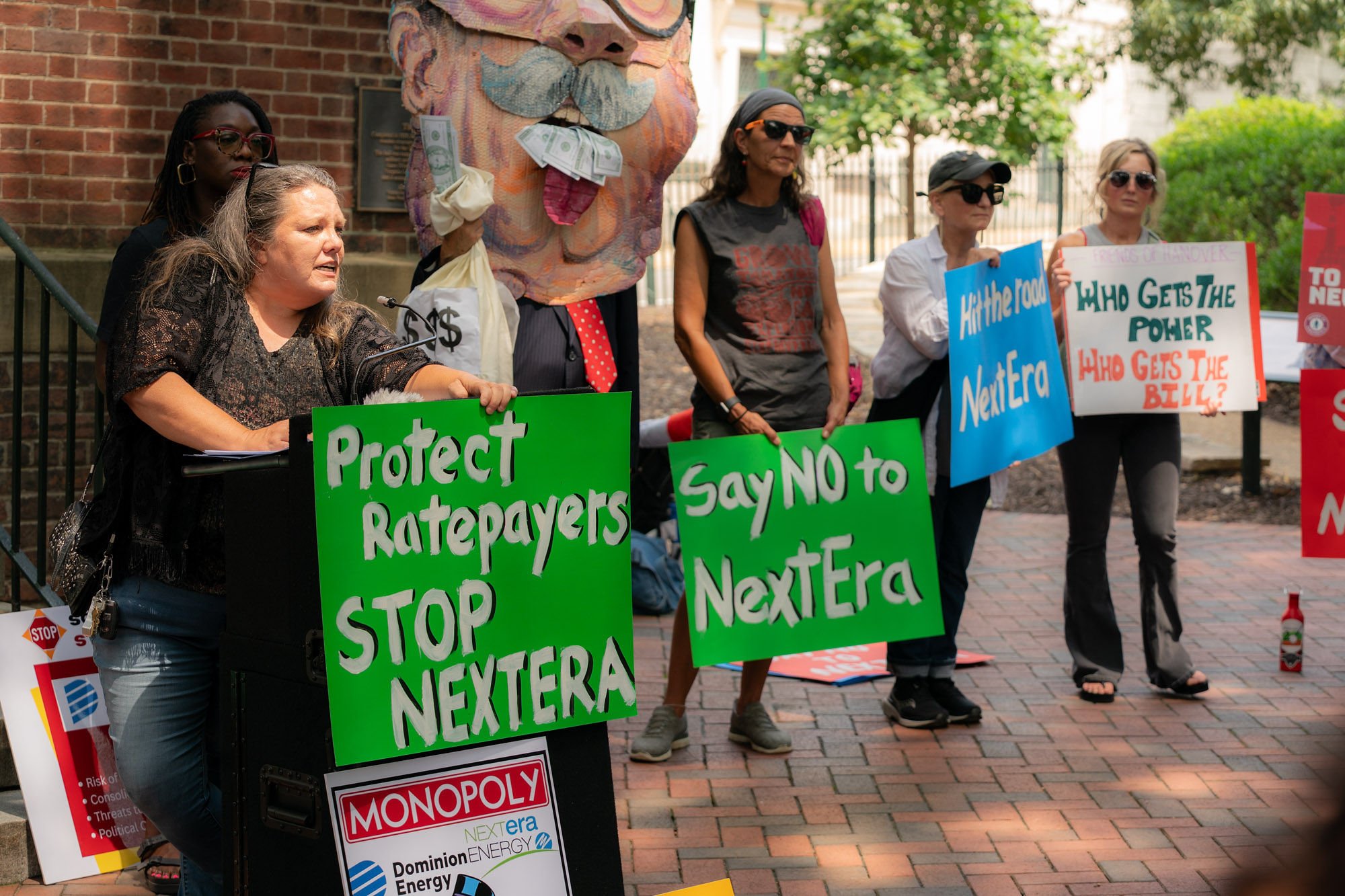 photo of woman speaking at podium with a sign in front of her that says protect ratepayers stop nextera, along with other people standing next to her with similar signs