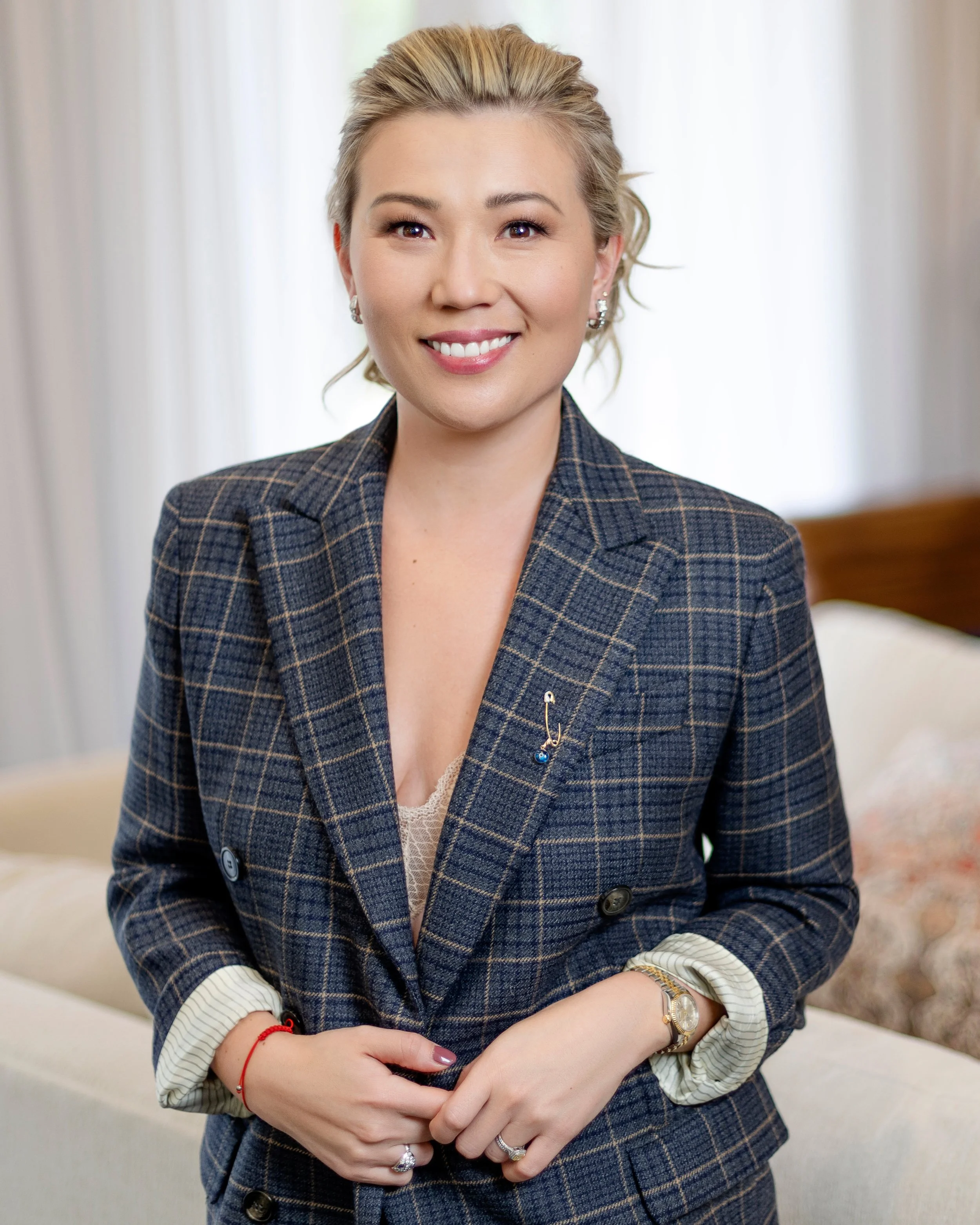 A woman with blonde hair smiling and wearing a plaid blazer, with her hands clasped in front of her. She has jewelry including earrings, rings, a watch, and a bracelet. A professional real estate sales agent.