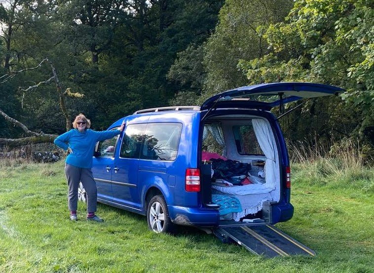 A woman in a blue hoodie and gray pants stands next to a blue camper van, which is equipped with a wheelchair-accessible ramp. The van is parked on grassy terrain with trees in the background. The rear hatch of the van is open, revealing a bed with blankets and luggage inside.