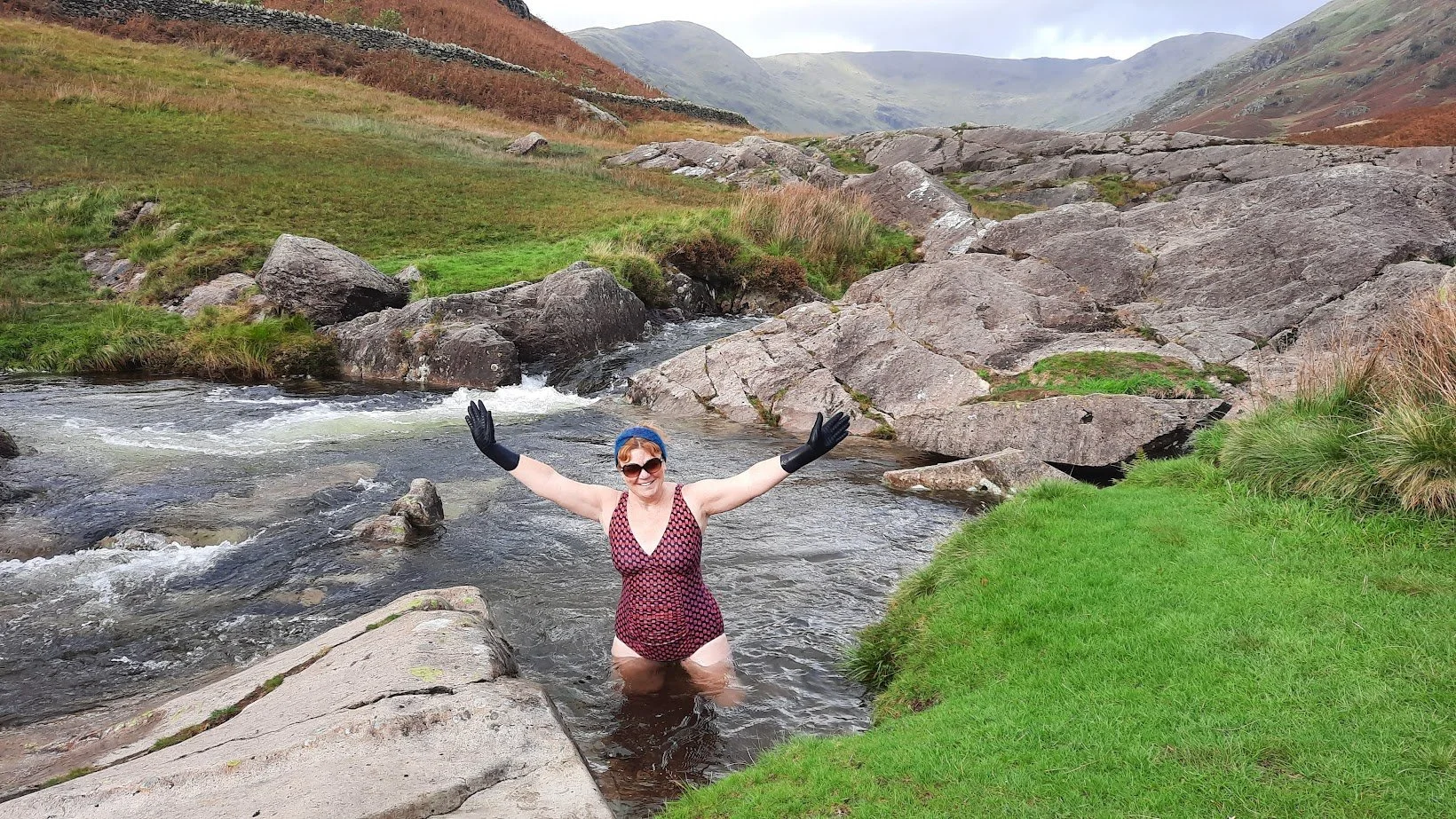 Woman standing in a river with outstretched arms, wearing sunglasses, a red and black swimsuit, black gloves, and a blue headscarf, in a lush green and rocky mountainous landscape.